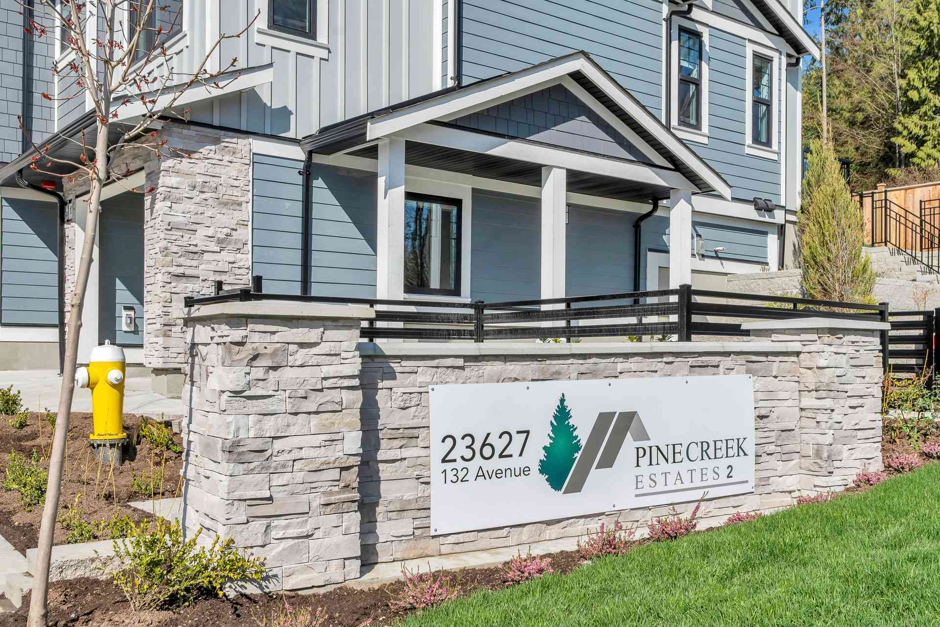 Pine Creek Estates – A ground-level entrance sign mounted on a stacked-stone low wall in front of a modern blue siding and stone-accented residential building. The shot features bright daylight, a green lawn, neat landscaping, a yellow fire hydrant, and black metal railings.