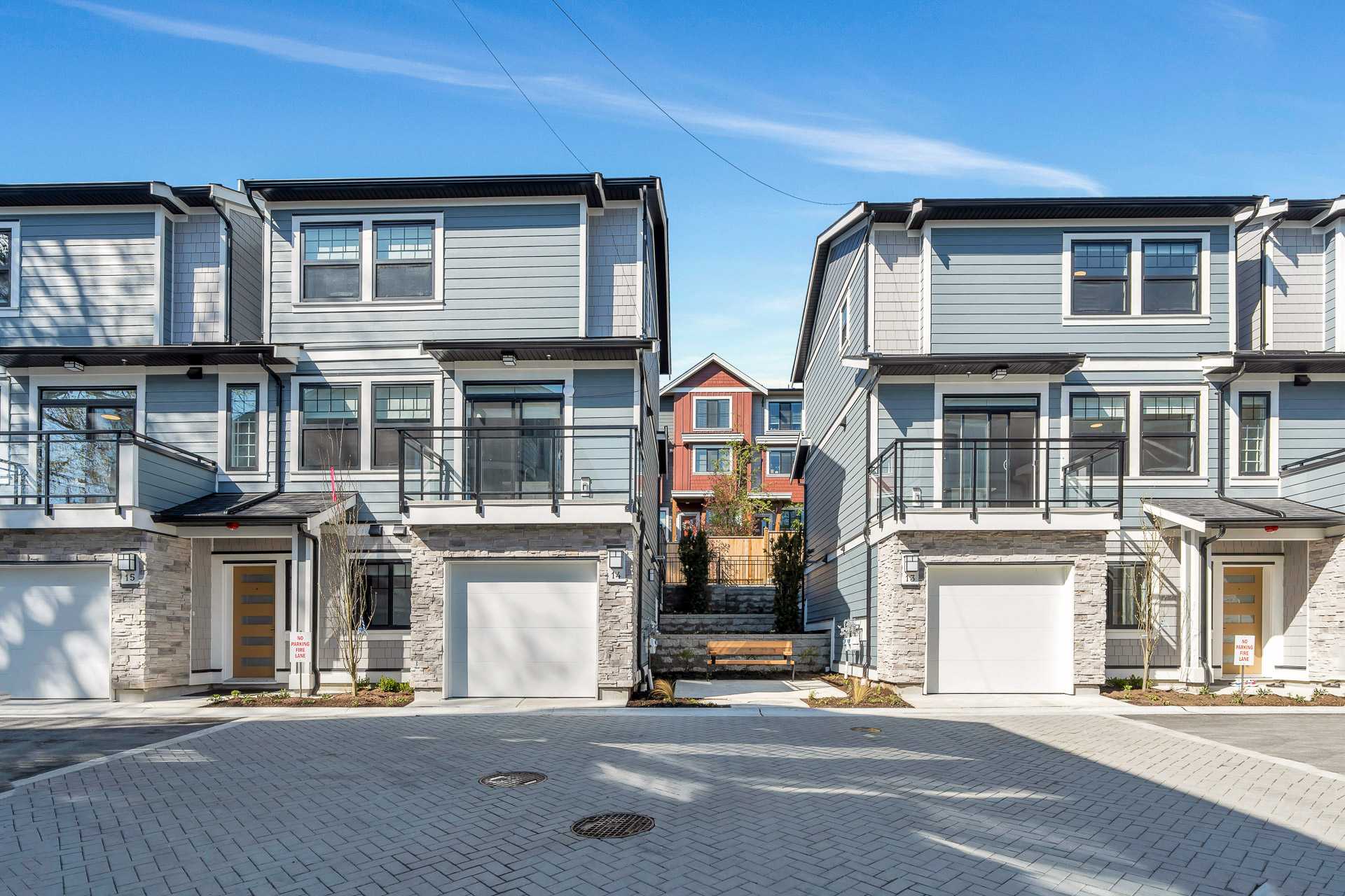 Pine Creek Estates – A ground-level street view of modern three-story townhomes with light blue lap siding, stacked stone accents, glass balcony railings, and single-car garages facing a paved courtyard. A small landscaped walkway with a wooden bench sits in the gap between the buildings under a bright blue, clear daytime sky.