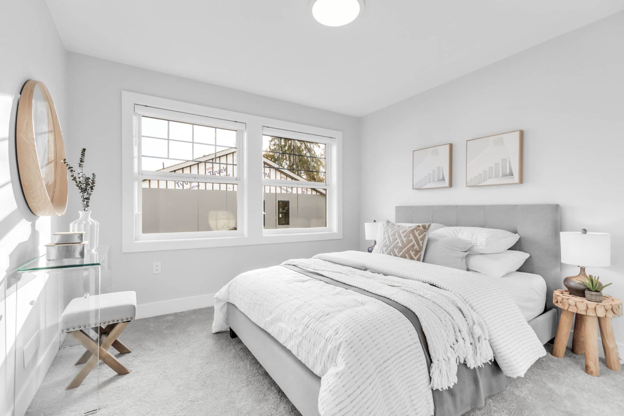 Pine Creek Estates – A modern bedroom features a upholstered grey bed with crisp white linens, light grey walls, and plush wall-to-wall carpeting. Sunlight streams through two large windows onto a clear acrylic vanity desk with a round wooden mirror and a matching padded stool.