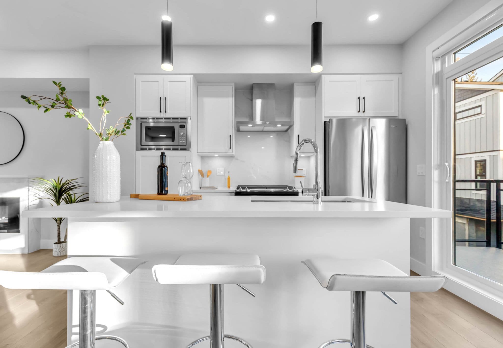 Pine Creek Estates – A modern, bright kitchen featuring a large white quartz island with three chrome barstools and two sleek black pendant lights hanging above. White cabinetry, stainless steel appliances, and a marble-patterned backsplash create a clean, contemporary aesthetic filled with natural daylight from an adjacent sliding glass door.