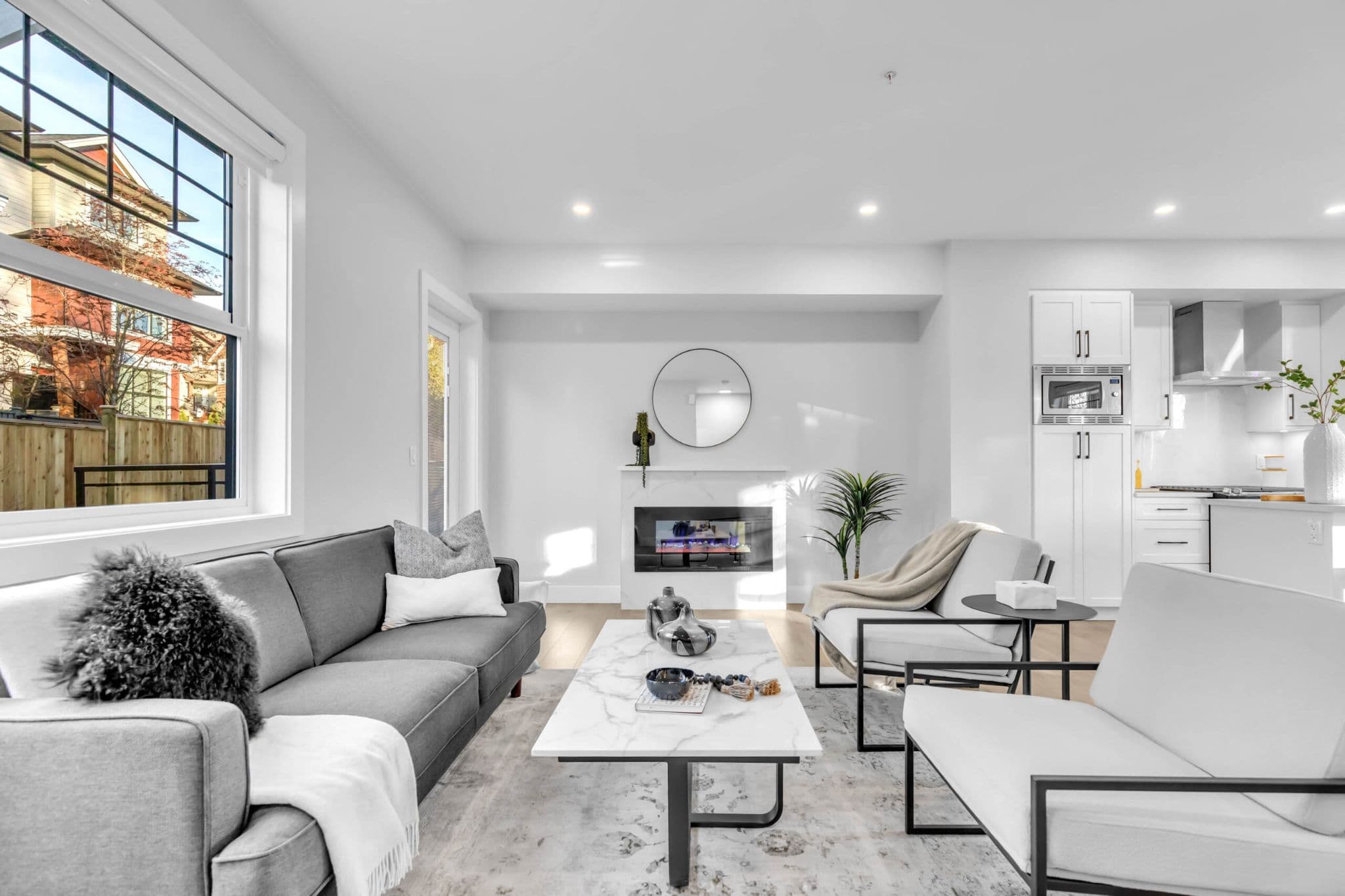 Pine Creek Estates – A bright, open-concept living room features a charcoal gray sofa, light gray armchairs, and a marble coffee table anchored by a modern linear fireplace with a round mirror above it. Natural daylight fills the space through a large window, revealing a clean white kitchen with sleek cabinetry in the background.