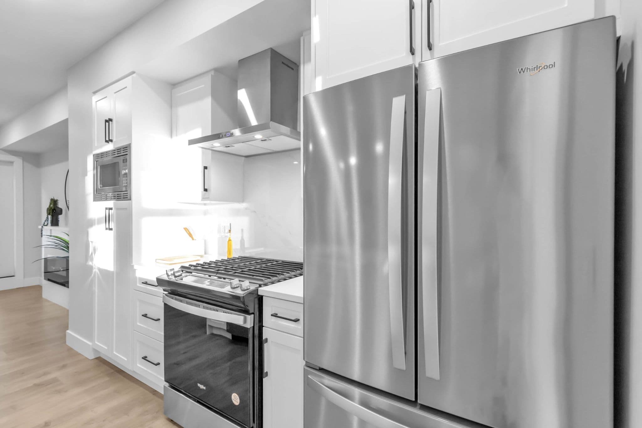 Pine Creek Estates – A modern kitchen featuring stainless steel Whirlpool appliances, including a French door refrigerator, gas range with a chimney hood fan, and a built-in microwave. Bright white shaker cabinetry, white countertops, and under-cabinet lighting create a clean, contemporary feel over light wood flooring.