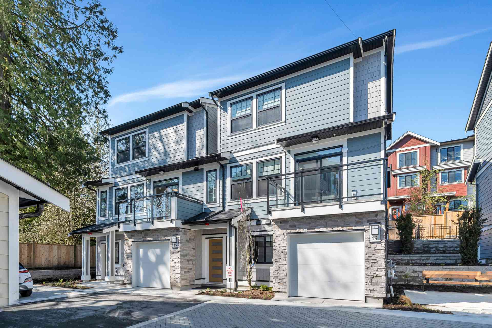 Pine Creek Estates – A ground-level view shows modern multi-story townhomes with light blue lap siding, white trim, stone masonry accent walls, and white garage doors under a clear blue sky. Each unit features glass-railed balconies on the upper floor, paved driveways, and small landscaped planting beds along the front entrances.