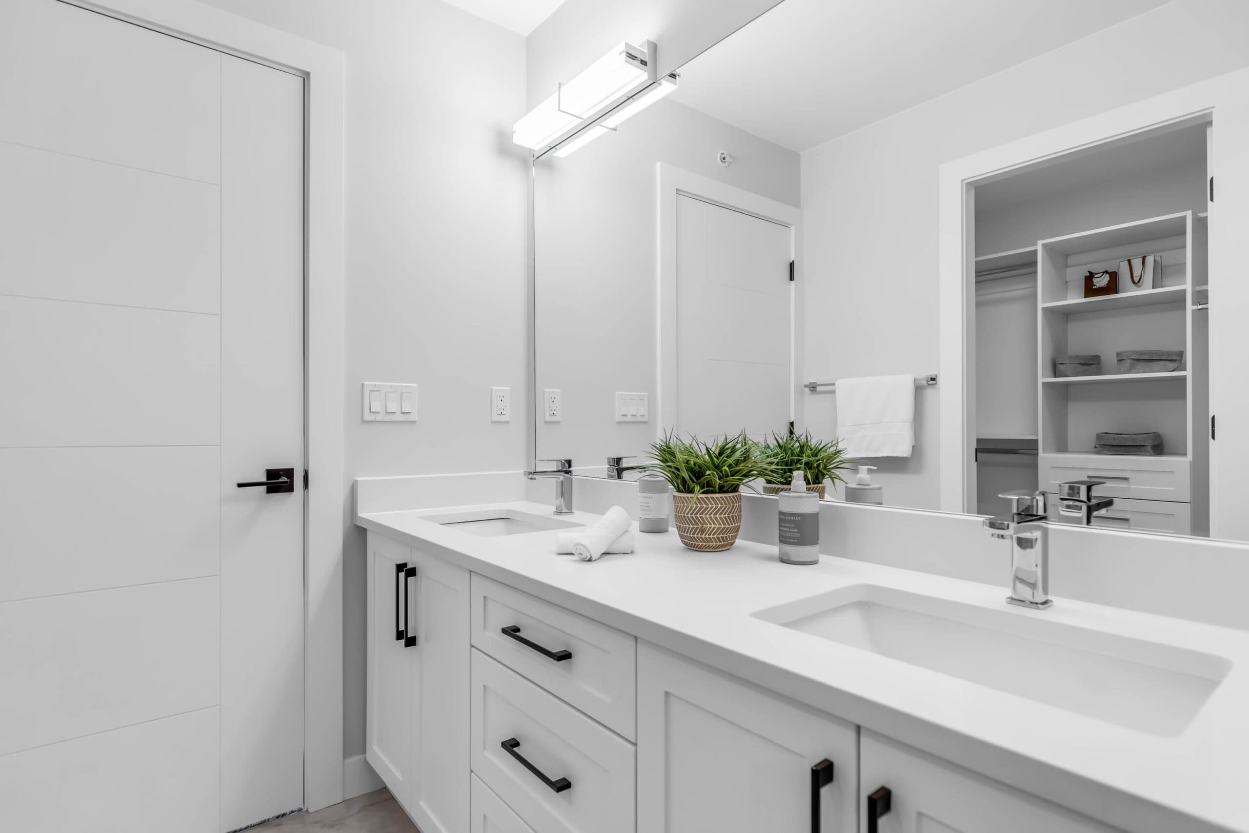 Pine Creek Estates – A modern, bright bathroom featuring a double-sink white vanity with matte black hardware and polished chrome faucets under a large wall-to-wall mirror. Bright vanity lighting illuminates clean white walls, potted faux greenery, and a reflection leading into an adjoining walk-in closet with built-in shelving.