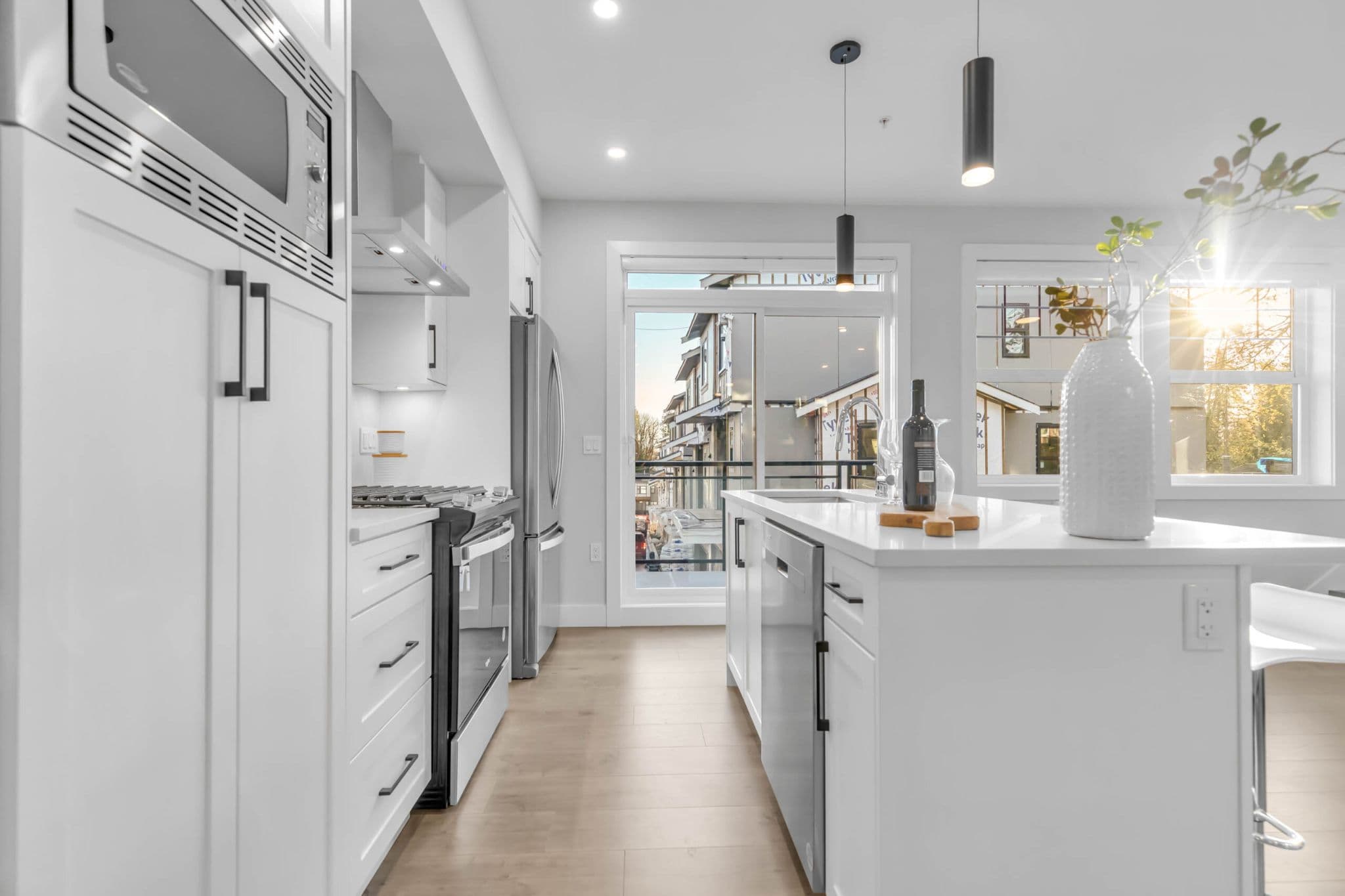 Pine Creek Estates – A modern, bright condo kitchen featuring white cabinetry, light hardwood flooring, and stainless steel appliances centered around a large kitchen island with quartz countertops. Sunlight streams through a large window and adjacent sliding glass balcony door, illuminating black hardware accents and sleek pendant lights.