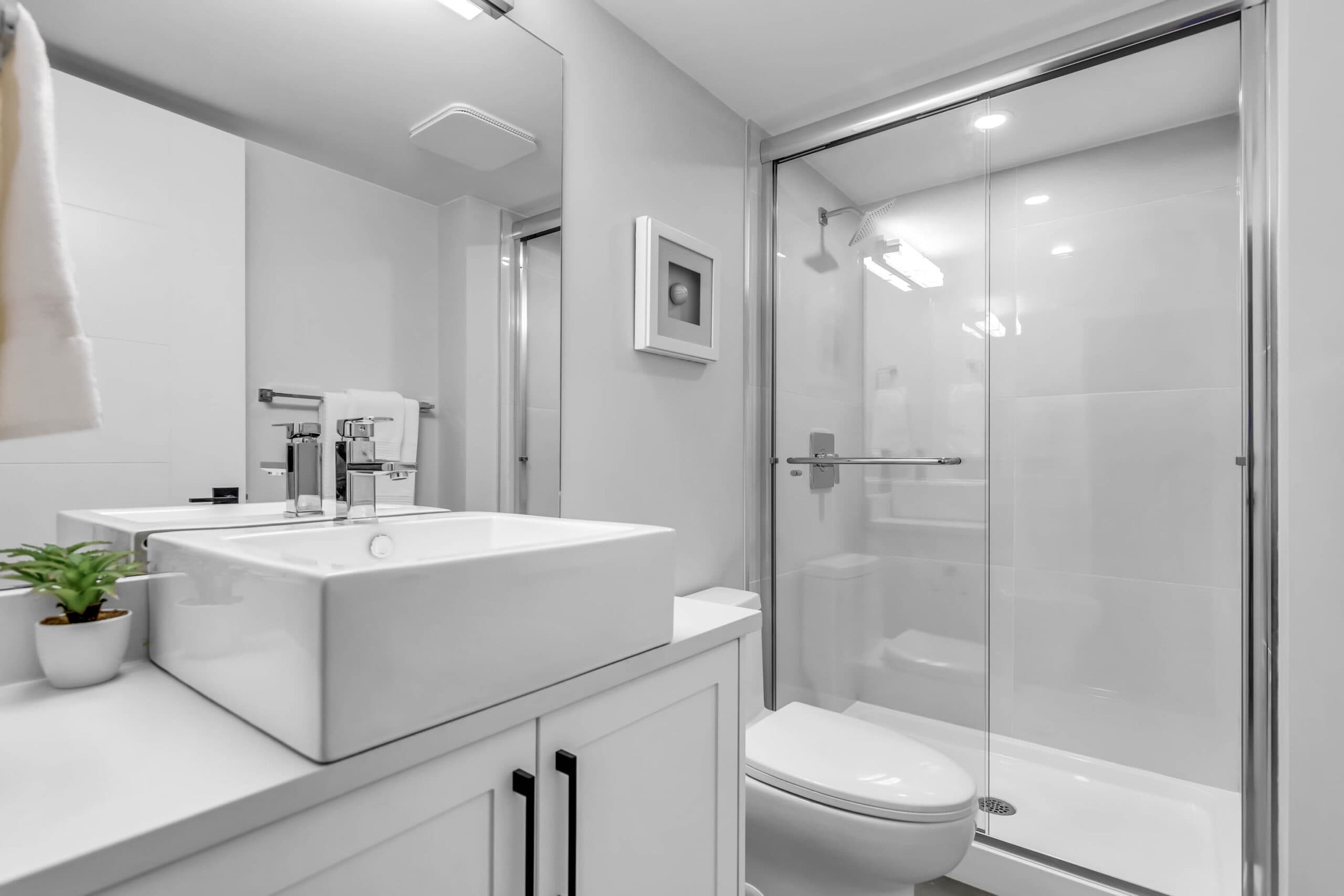 Pine Creek Estates – A modern, bright bathroom featuring a white vessel sink atop a vanity cabinet with matte black hardware, positioned beside a flush-mount toilet. A glass-enclosed walk-in shower with white tile walls and chrome fixtures sits in the corner under soft recessed lighting.