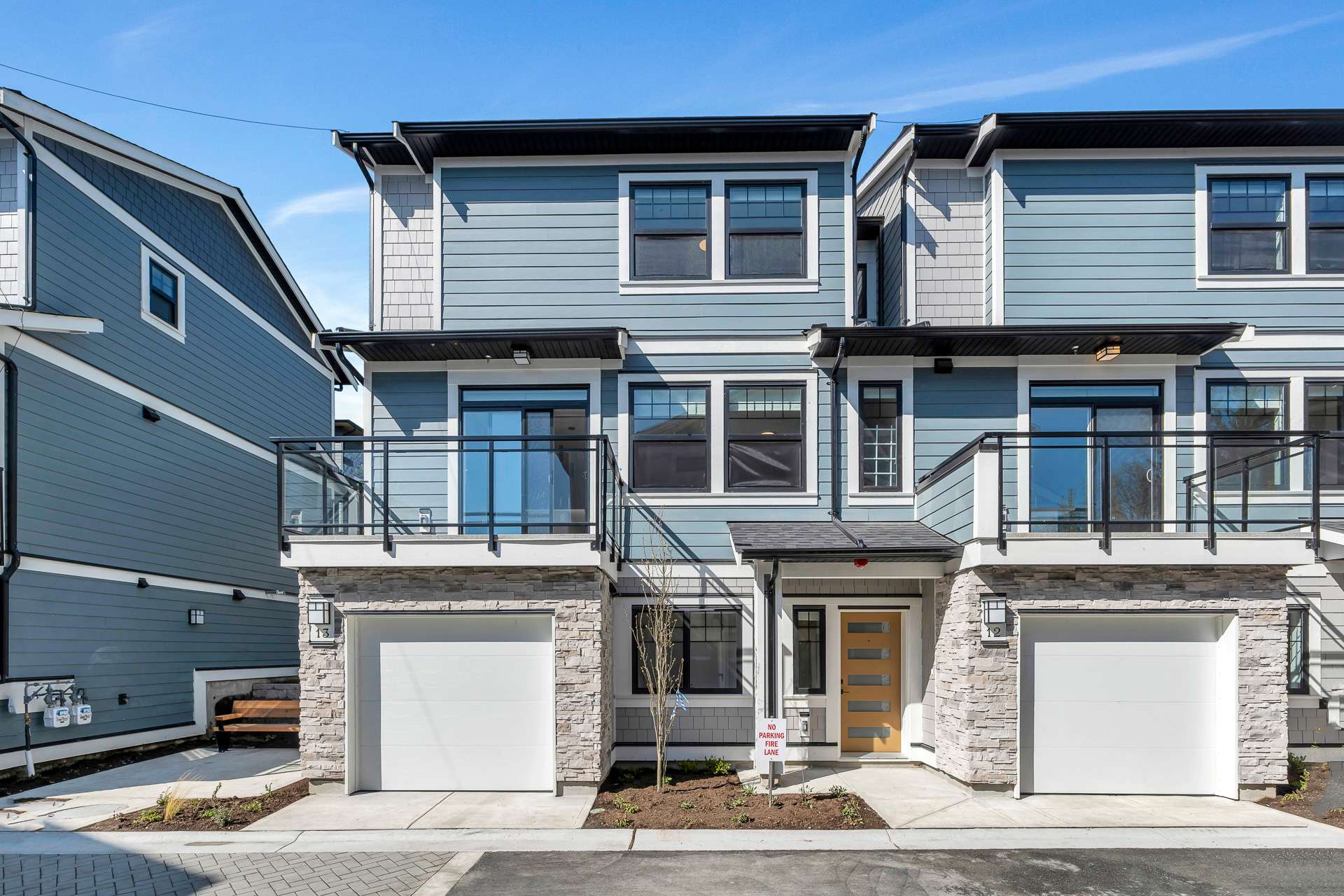 Pine Creek Estates – Ground-level view of modern three-story townhomes featuring light blue horizontal siding, grey shingle accents, stone-veneered lower levels, and white garage doors. Each unit includes a second-floor balcony with glass railings and a wooden front door under a bright, clear blue sky.