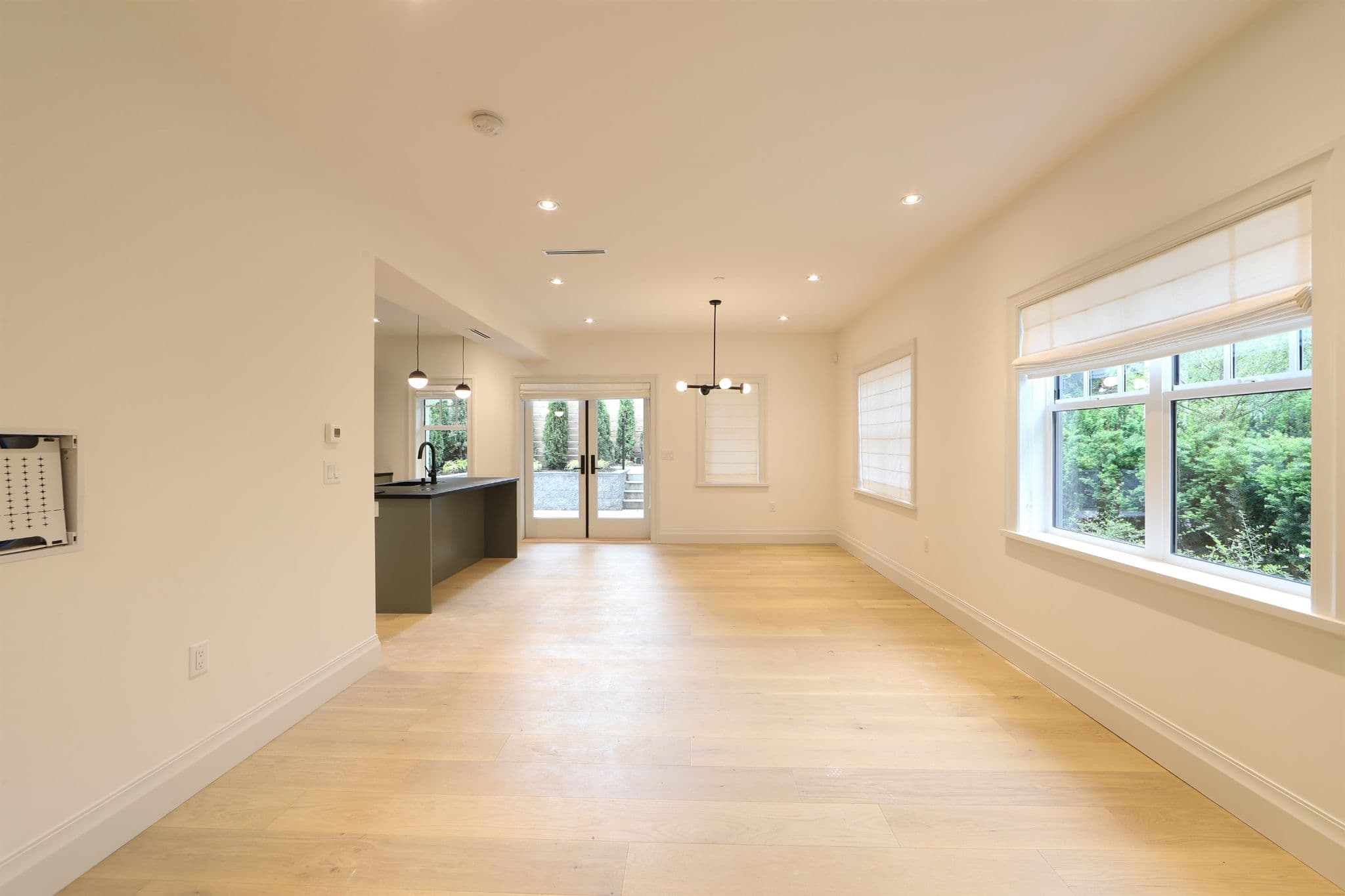 BLVD on the Greenway – A wide, vacant open-concept living and dining area featuring light-toned hardwood floors, crisp off-white walls, and recessed ceiling lighting. Large windows with roman shades frame views of lush outdoor greenery, leading toward a dark kitchen island and double glass doors opening to an exterior patio.