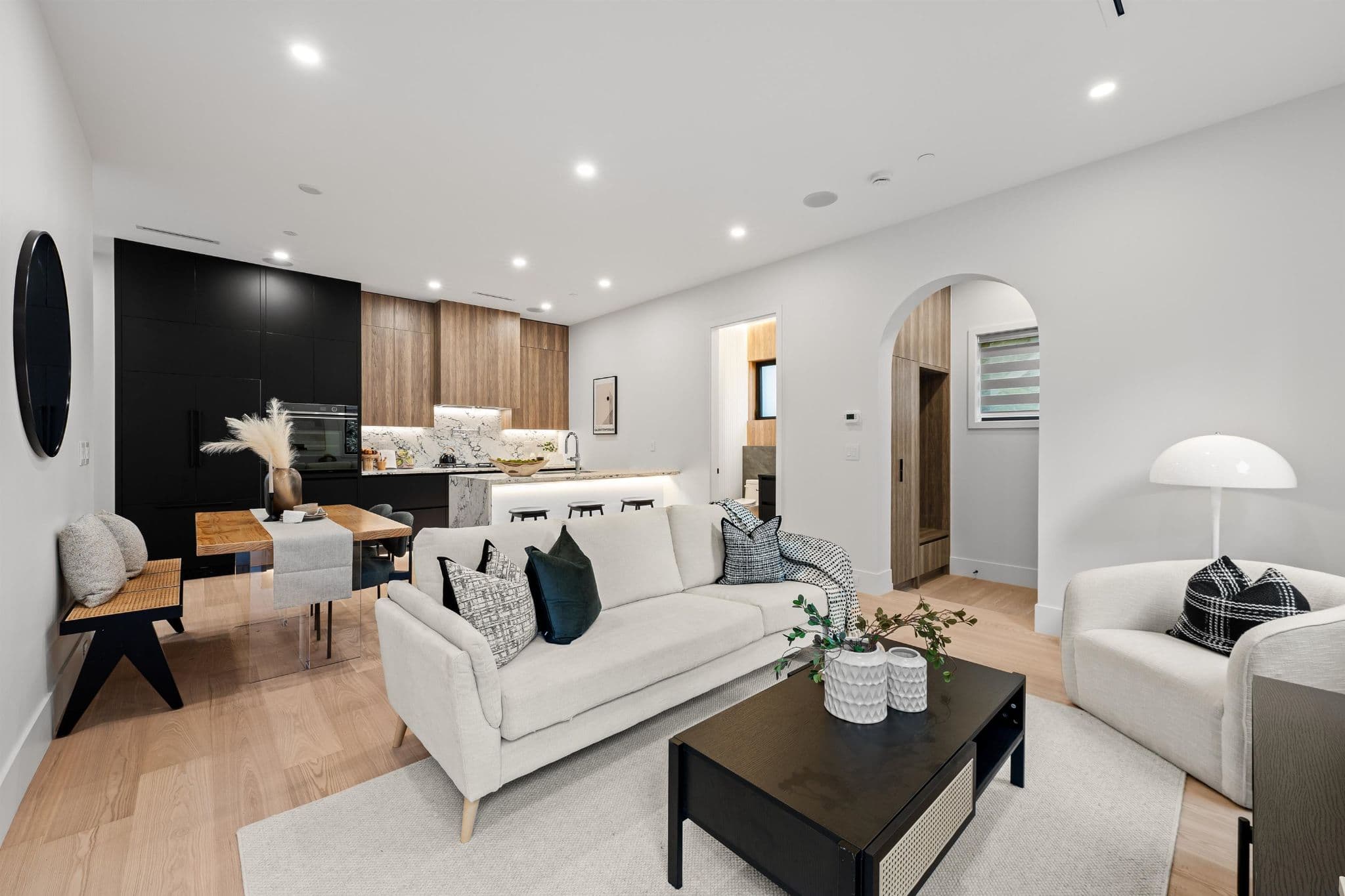 BLVD on the Greenway – A modern open-concept living, dining, and kitchen area featuring light hardwood floors and white walls with recessed ceiling lighting. The space is styled with a light fabric sofa, dark coffee table, and an accent armchair in the foreground, leading back to a wood-and-glass dining table and a kitchen with matte black and natural wood cabinetry, marble countertops, and an arched hallway opening.