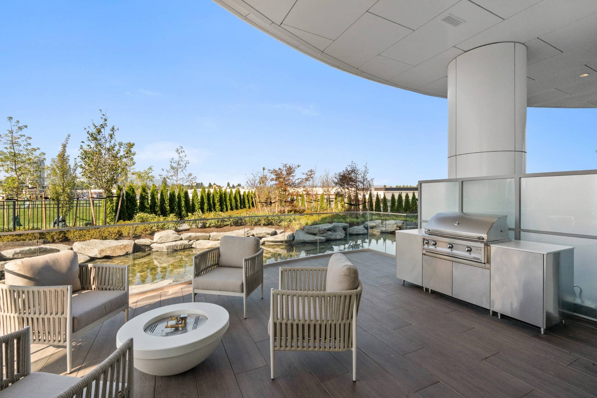 Icon at Southgate City – A covered outdoor terrace amenity equipped with a stainless steel BBQ cooking station, a round modern fire pit table, and woven lounge armchairs on dark tiled flooring. Glass railings overlook an adjacent landscaped rock pond garden and lush greenery under a clear daytime sky.