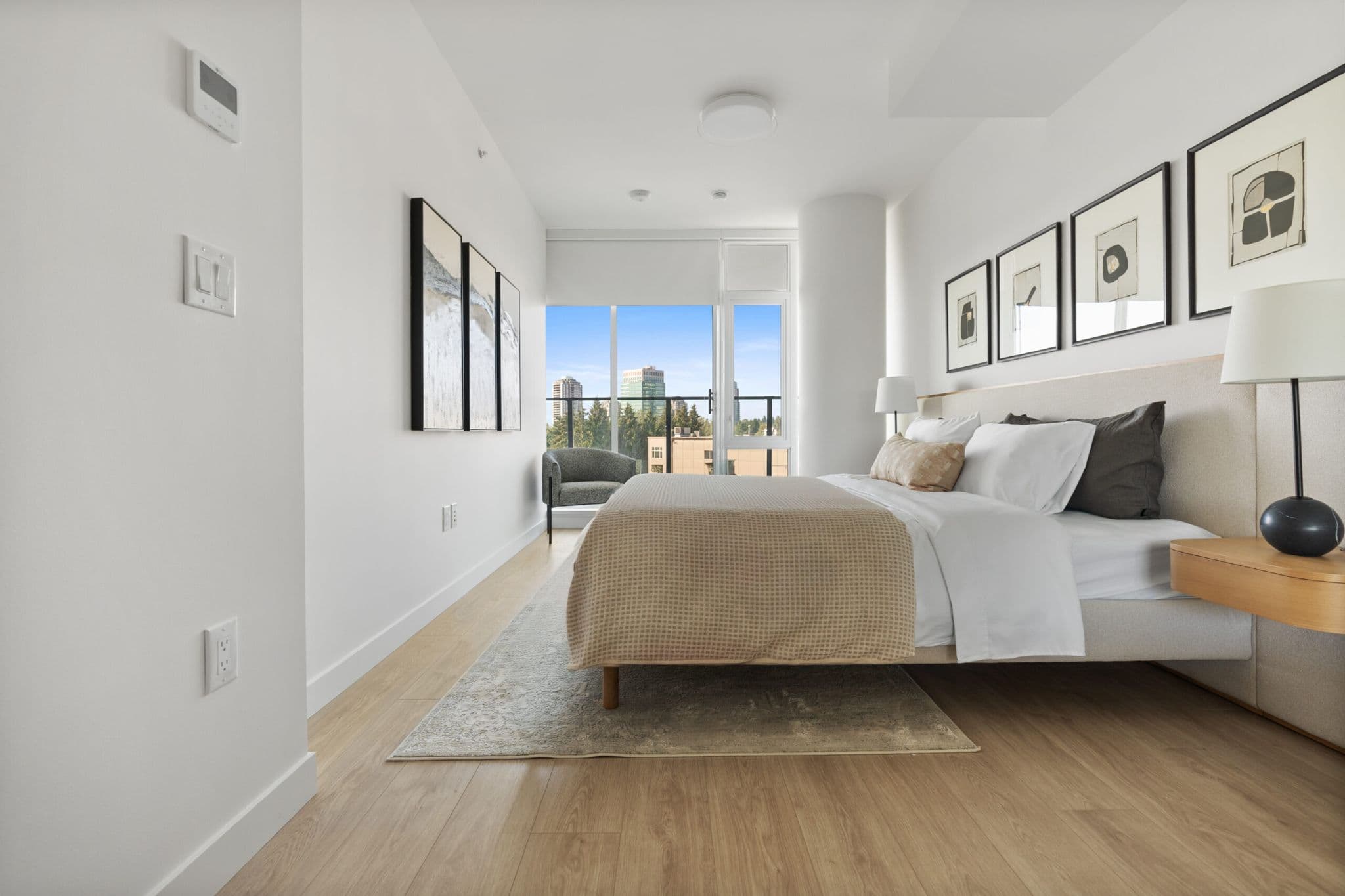 Icon at Southgate City – A bright, modern bedroom featuring light hardwood floors, crisp white walls decorated with framed artwork, and a neatly made bed with neutral-toned linens. At the far end, floor-to-ceiling windows and a glass door open to a balcony overlooking treetops and distant city high-rises under a clear blue daytime sky.