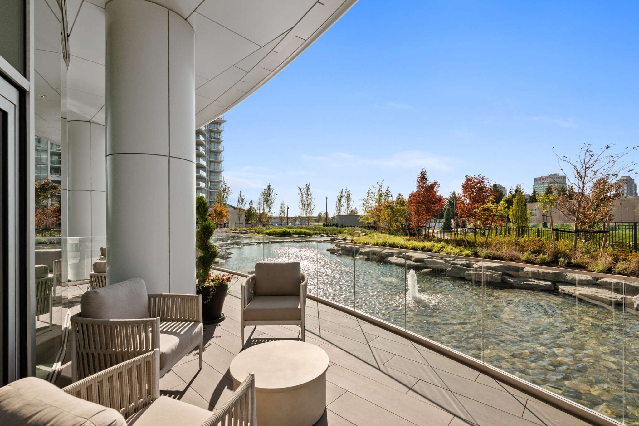 Icon at Southgate City – A covered outdoor terrace featuring modern lounge armchairs and a low table sits behind a clear glass railing overlooking a rocky water feature with fountain jets. Landscaped grounds with colorful autumn trees and neighboring residential towers extend under a bright blue, sunlit sky.