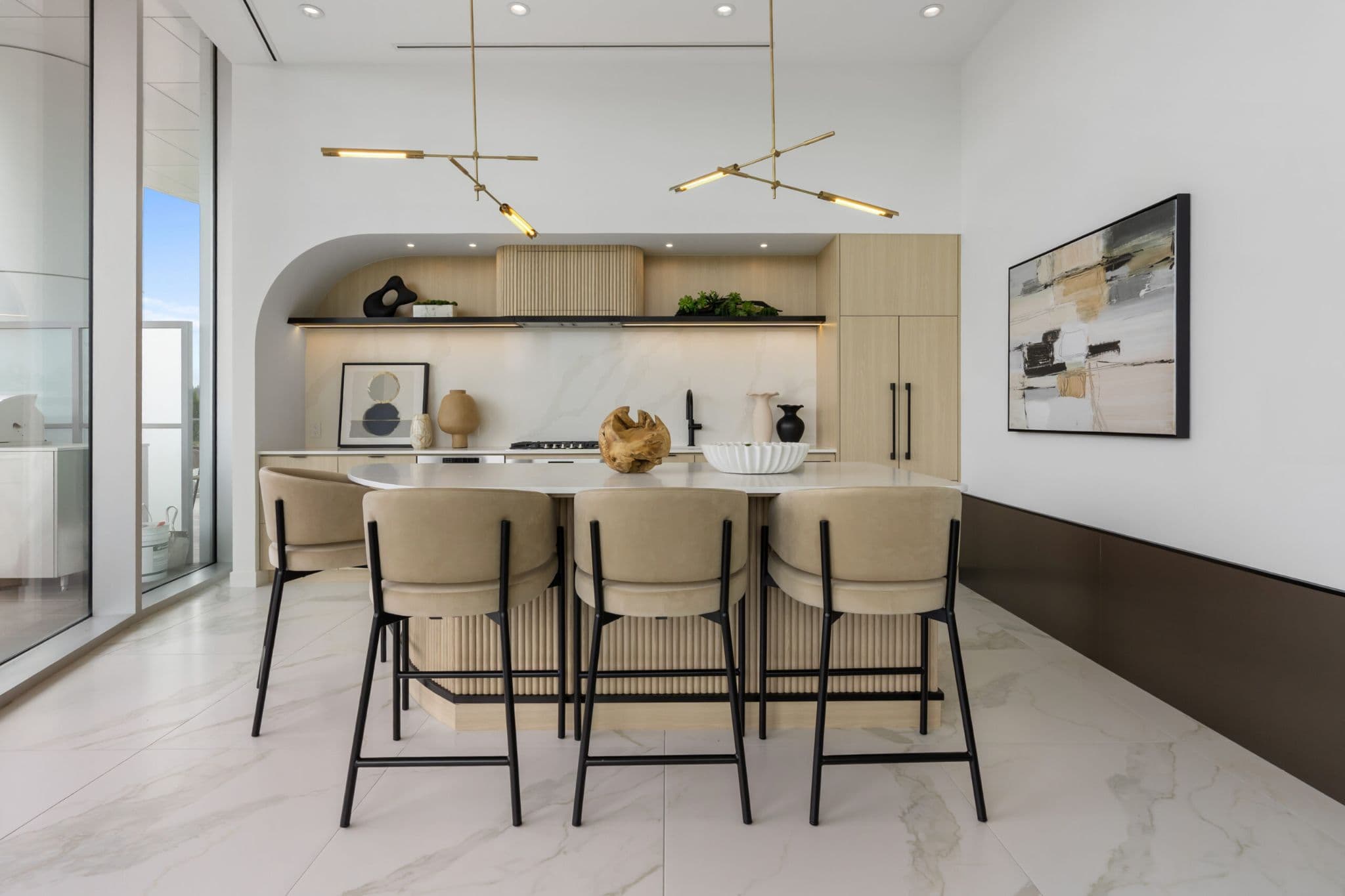 Icon at Southgate City – A bright, modern kitchen featuring an arched alcove with light wood cabinetry, a fluted range hood, and marble countertops and backsplashes. In the foreground, four neutral upholstered barstools sit at a rounded, fluted island beneath sculptural brass pendant lights, with floor-to-ceiling glass doors opening onto an outdoor balcony.