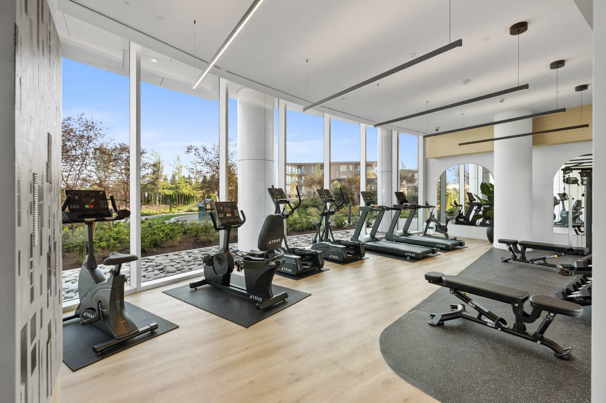 Icon at Southgate City – A bright, modern fitness center features a line of stationary bikes, ellipticals, and treadmills facing floor-to-ceiling windows with views of landscaped gardens and trees. The space is equipped with light wood flooring, a dedicated dark rubber workout area with weight benches, arched mirrors, and minimalist linear ceiling lights.