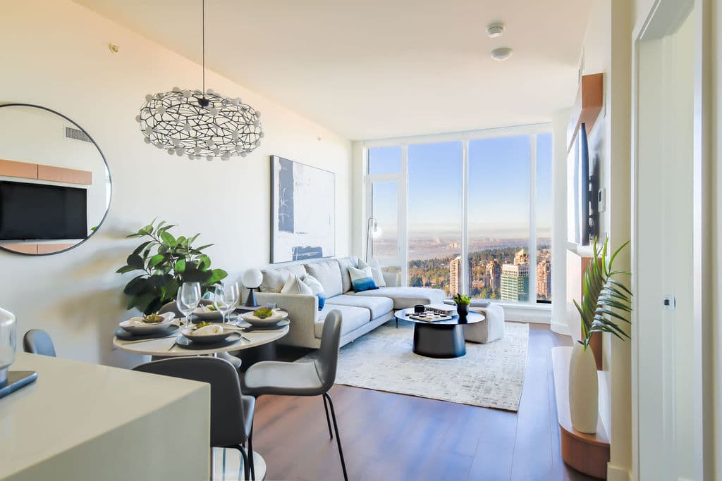 Icon at Southgate City – A bright, open-concept high-rise living and dining area featuring hardwood floors, a round dining table set for four under a sculptural wire light fixture, and a light gray sectional sofa. Floor-to-ceiling windows at the far end flood the modern space with daylight and frame sweeping panoramic views of city towers, forests, and distant water under a clear blue sky.