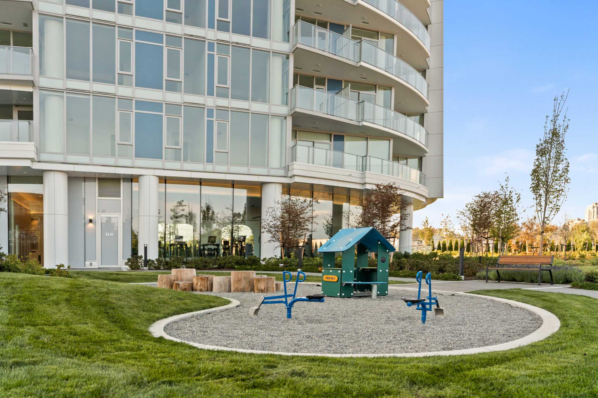 Icon at Southgate City – A ground-level outdoor children's playground featuring a small playhouse, blue diggers, and log stools set within a gravel play pit and green lawn. Behind the play area stands a modern curved high-rise residential building with floor-to-ceiling glass windows and private balconies.