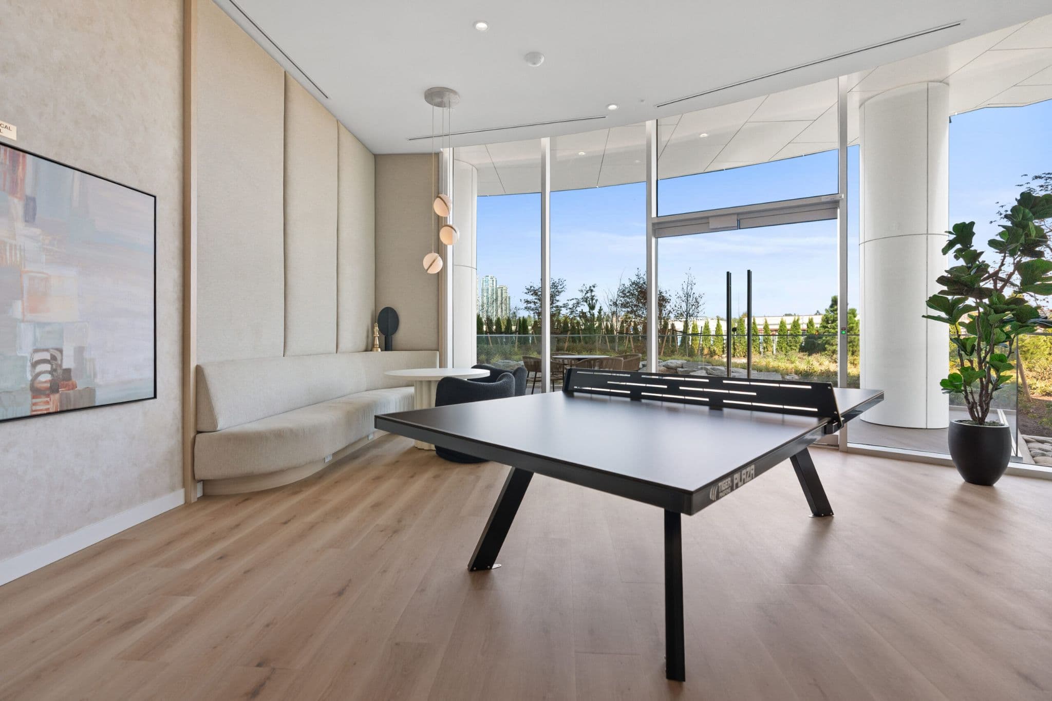 Icon at Southgate City – A bright indoor games and lounge amenity room featuring a black ping pong table centered on light wood floors. Floor-to-ceiling windows look out onto an outdoor terrace and landscaped greenery, complemented by a curved upholstered banquette, small seating nook, and modern pendant lighting.