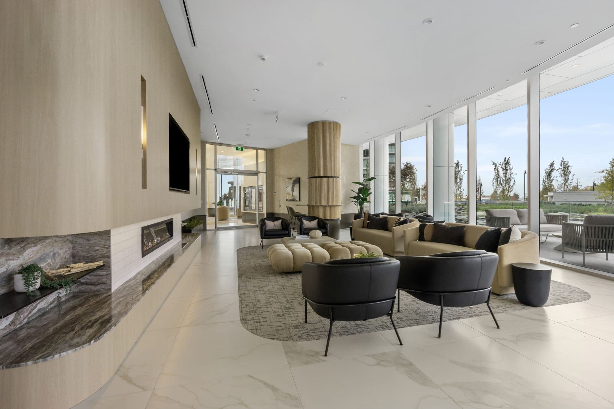 Icon at Southgate City – A bright and spacious contemporary residential lounge featuring light-wood paneled accent walls, a linear electric fireplace, and plush seating arranged with sofas, armchairs, and a large tufted ottoman. Floor-to-ceiling windows look out onto an outdoor patio and landscaped greenery under natural daylight.