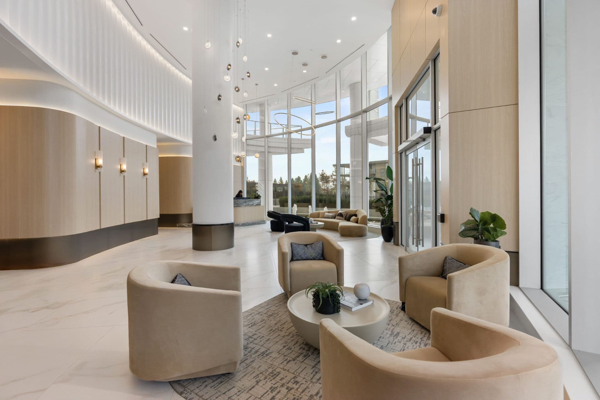 Icon at Southgate City – A bright, high-ceilinged modern lobby featuring large floor-to-ceiling glass walls, warm wood-paneled accents, and marble tile flooring. Contemporary seating areas with plush beige armchairs and curved sofas are arranged throughout the space beneath artistic pendant lighting and near a concierge reception desk.