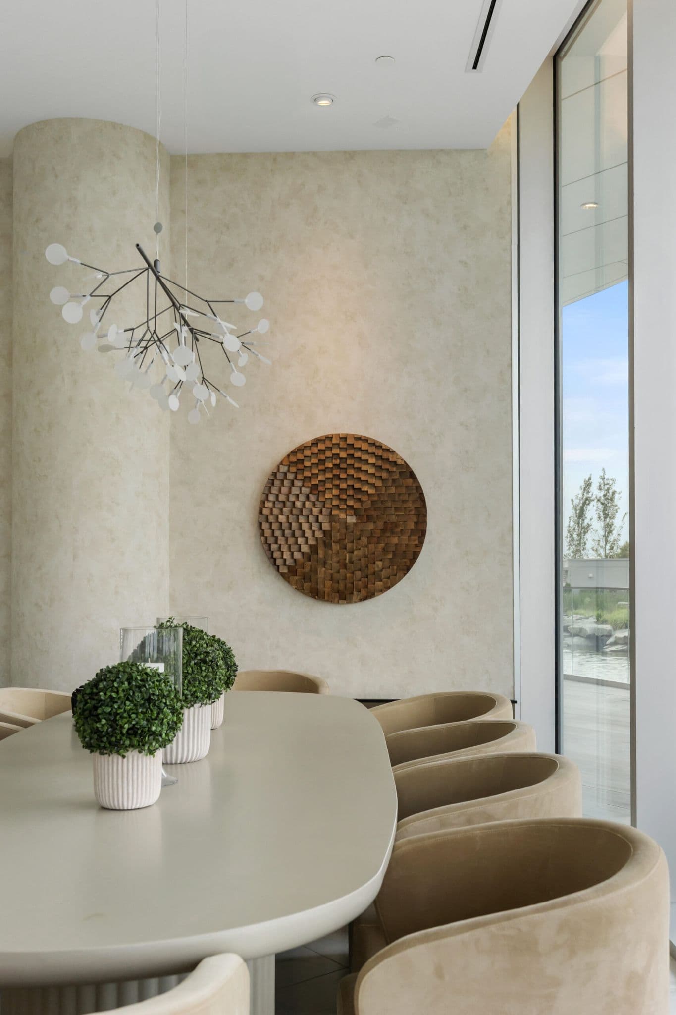 Icon at Southgate City – A modern conference or dining area with a long oval table surrounded by beige curved velvet armchairs, set against textured cream walls with a circular wooden art piece and a contemporary disc chandelier. Floor-to-ceiling windows on the right let in natural daylight and offer views of an outdoor water feature and greenery.