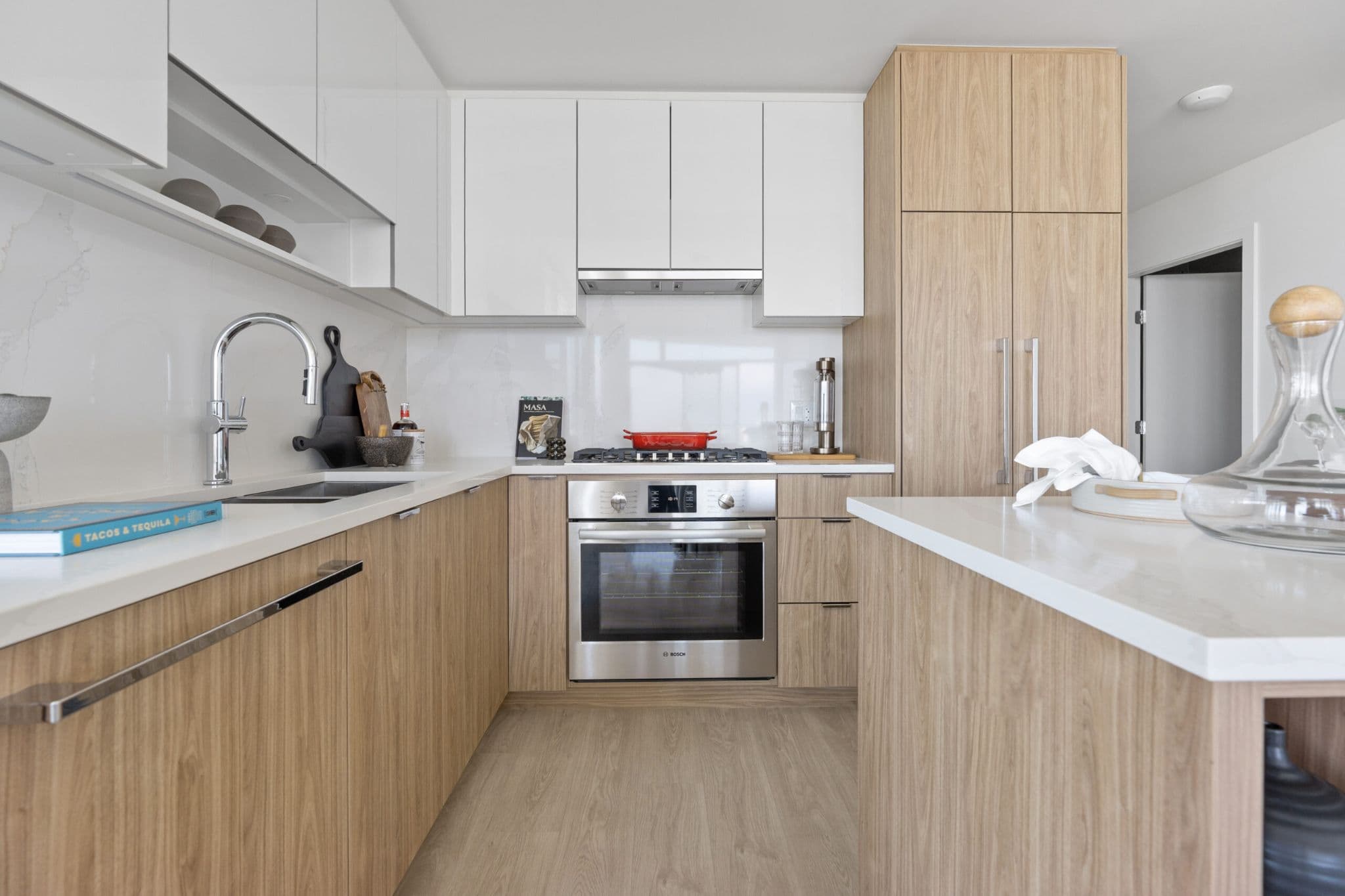 Icon at Southgate City – A modern, bright kitchen features two-tone cabinetry with light natural wood lower units and glossy white upper cabinets against a white marble backsplash. Integrated stainless steel appliances, white solid countertops, and light wood flooring complete the clean, contemporary aesthetic.