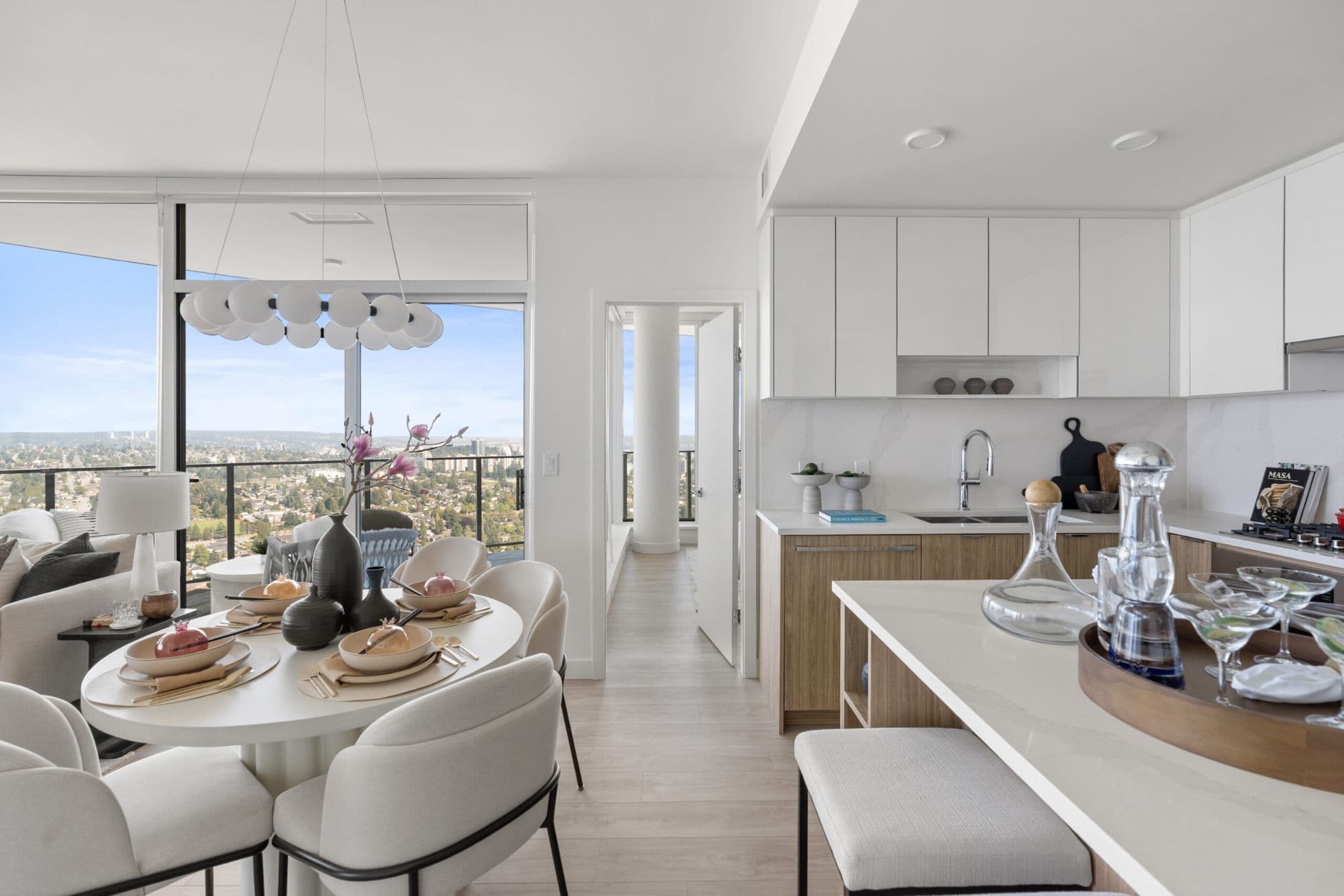 Icon at Southgate City – A bright, open-concept high-rise kitchen and dining area featuring flat-panel white and wood cabinetry, a quartz island, and a set round dining table beneath a modern chandelier. Large floor-to-ceiling windows and a glass balcony door showcase an expansive panoramic city view under a clear daytime sky.