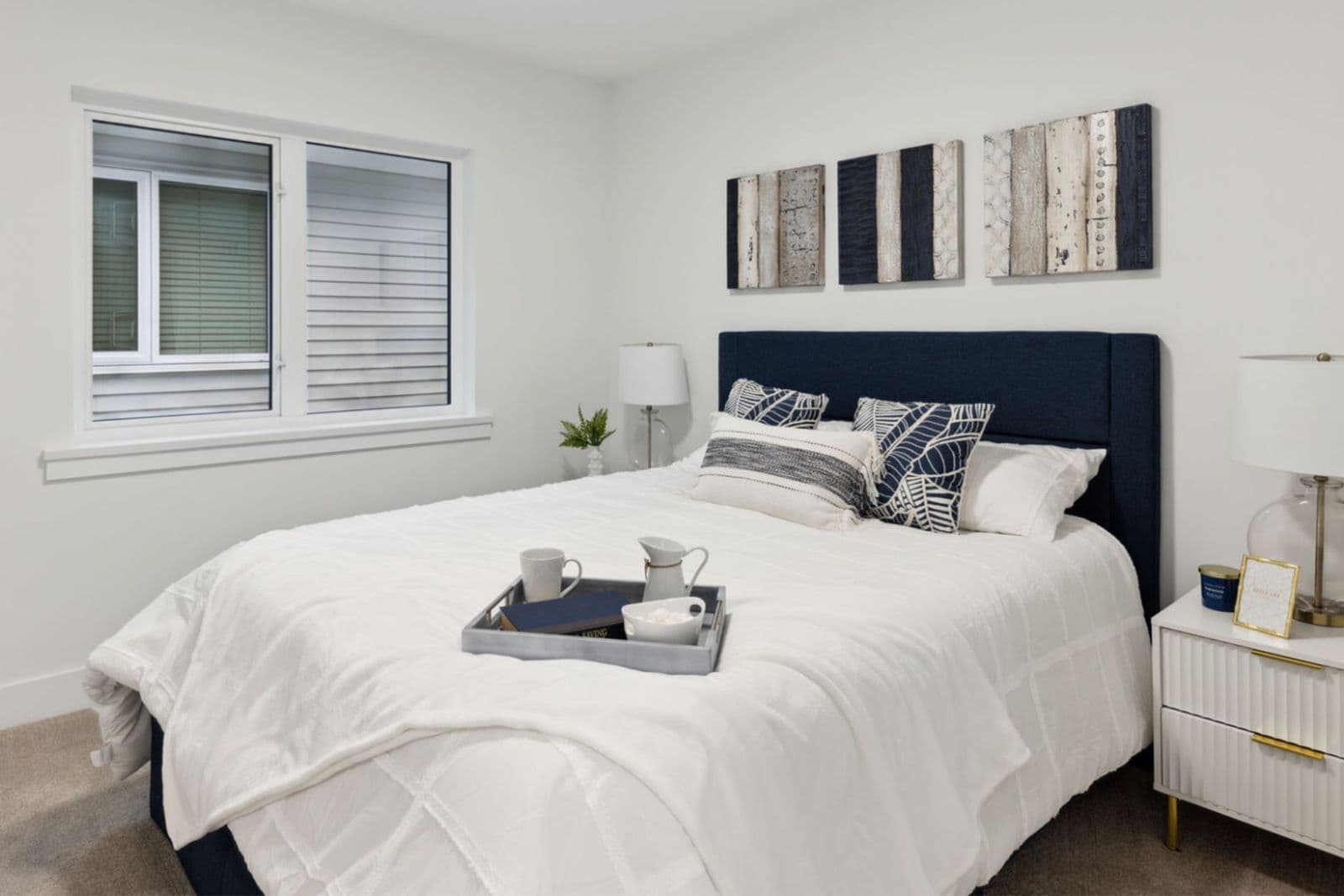 Brighton North – Bedroom with a dark blue upholstered headboard, white bedding, nightstand, and window.