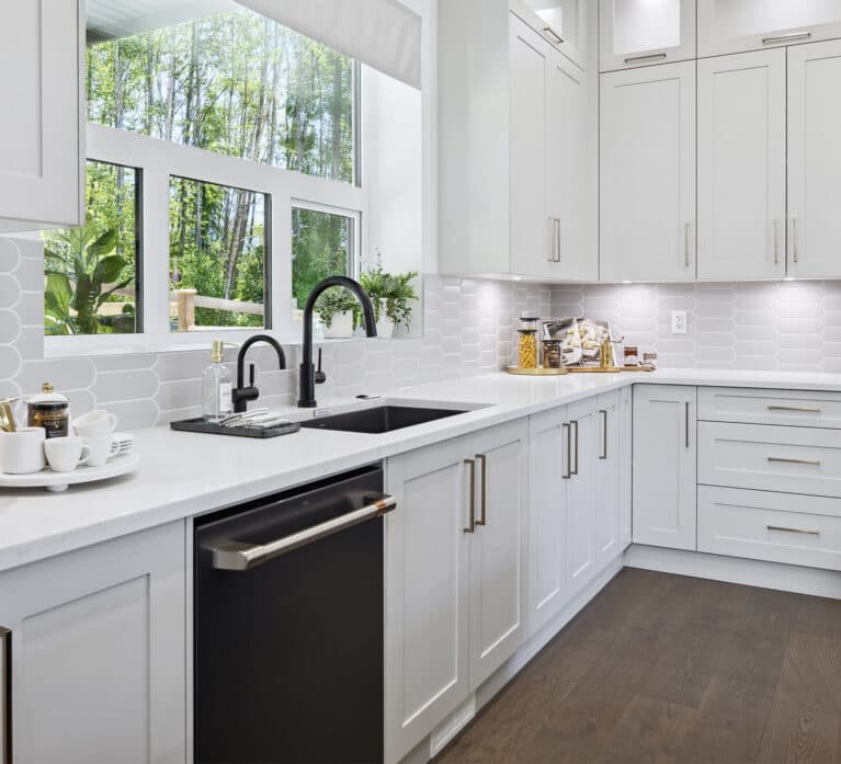 Brighton North – Close-up of kitchen perimeter counter with black sink, matte black faucet, dishwasher, white tiled backsplash, and white cabinetry.