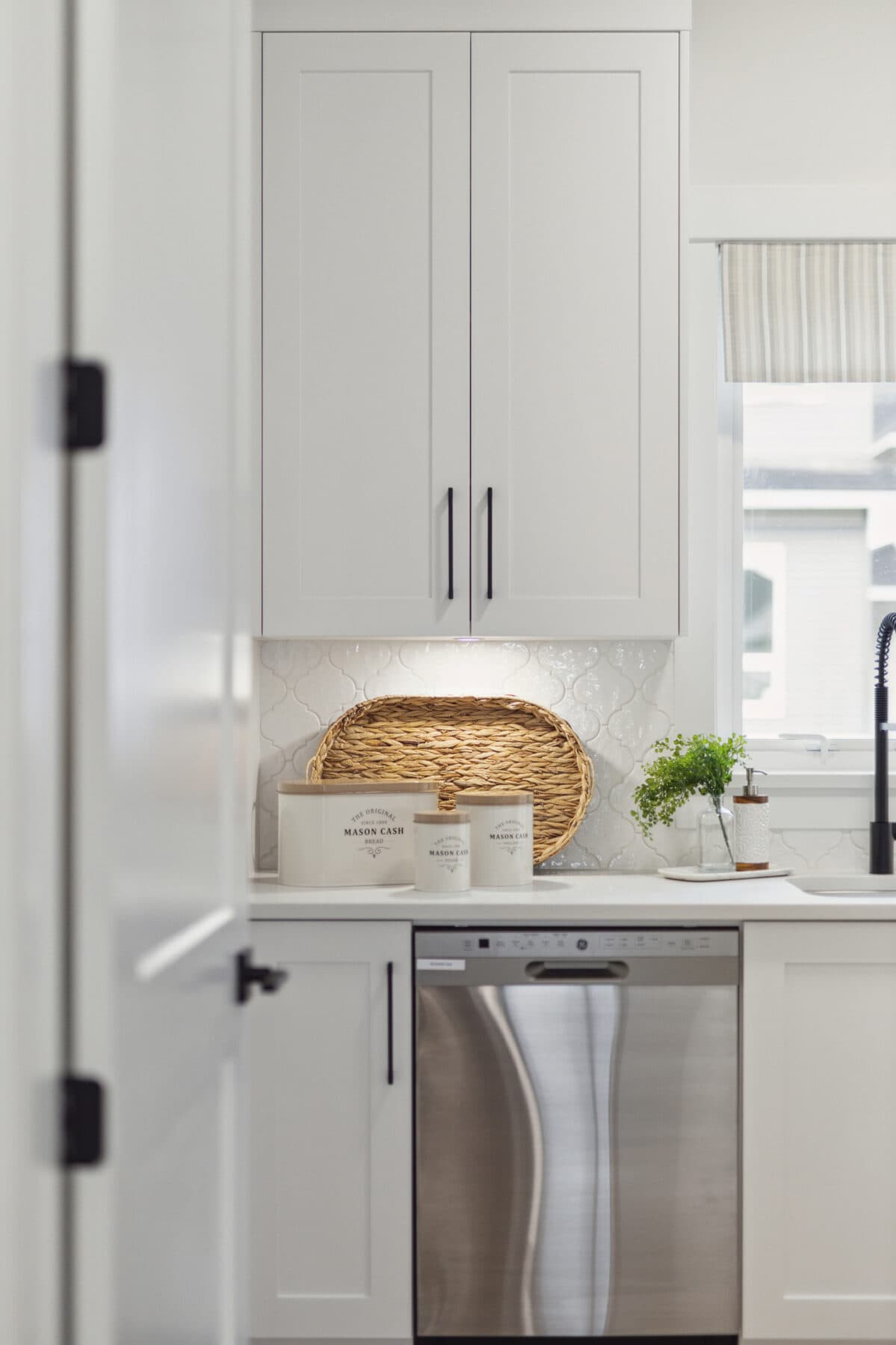 Brighton North – Kitchen detail highlighting white shaker cabinets, ceramic backsplash, countertop storage containers, and stainless steel dishwasher.