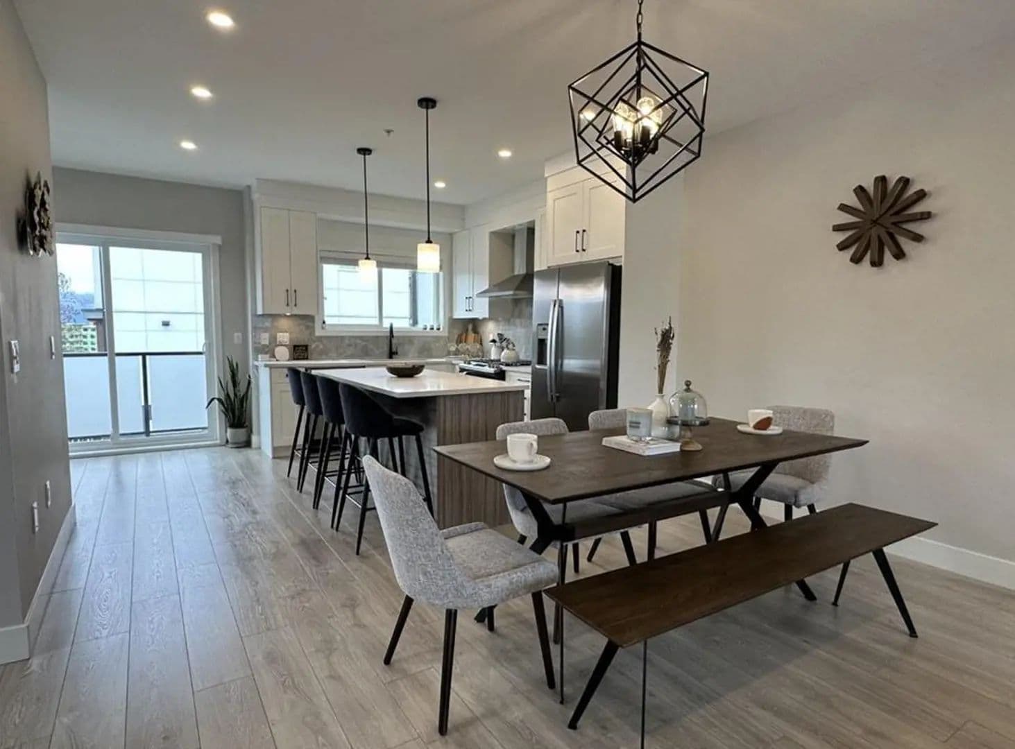 Willow Living – Open-concept kitchen and dining area with white cabinets, a stainless fridge, a dark wood dining table with bench seating, and a geometric pendant light.