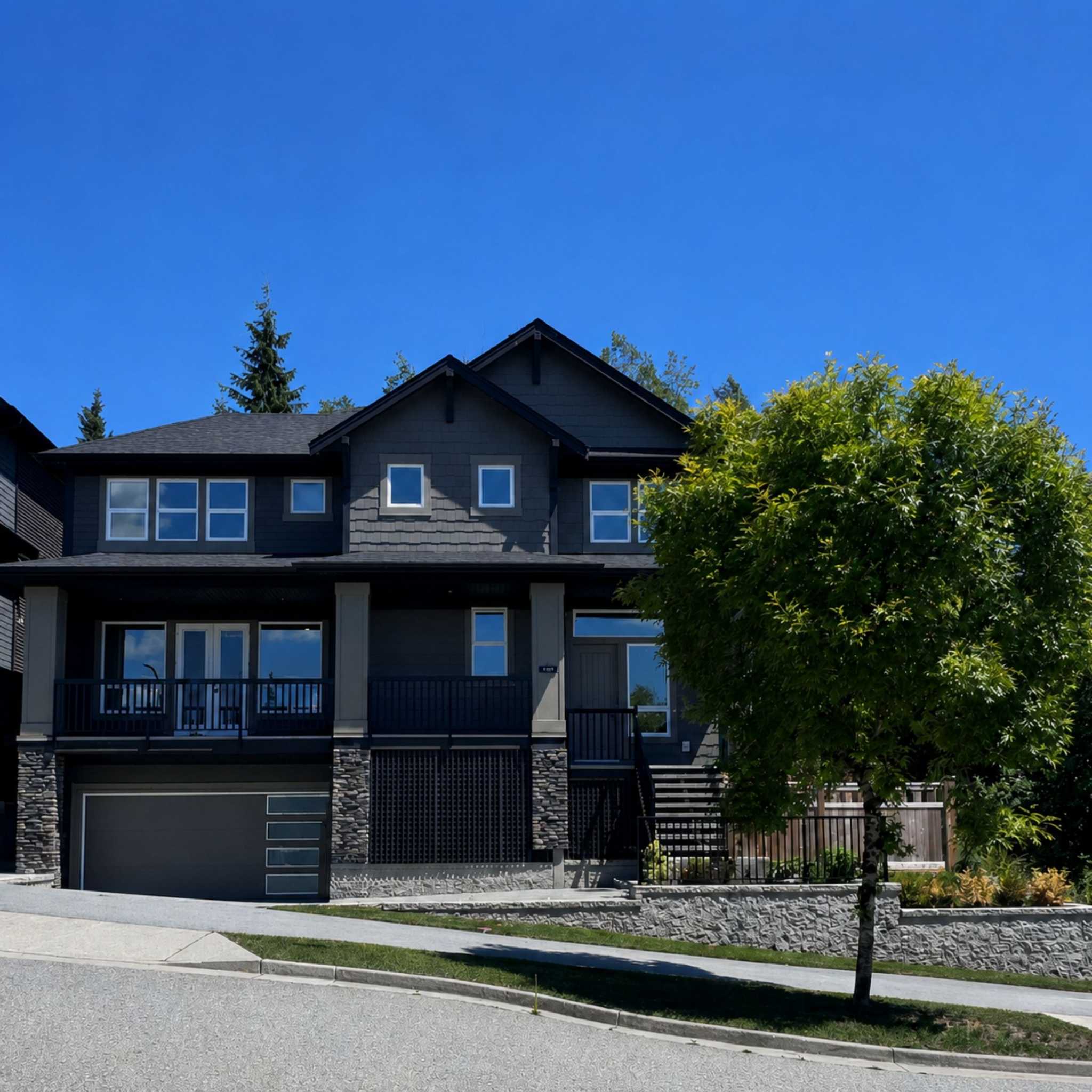 Harper Peak – A modern two-story dark charcoal house with stone accent pillars, a two-car garage, and a wide front porch sits on a sloping street under a clear blue sky. A leafy green tree stands in the manicured front yard alongside stone retaining walls and a paved driveway.