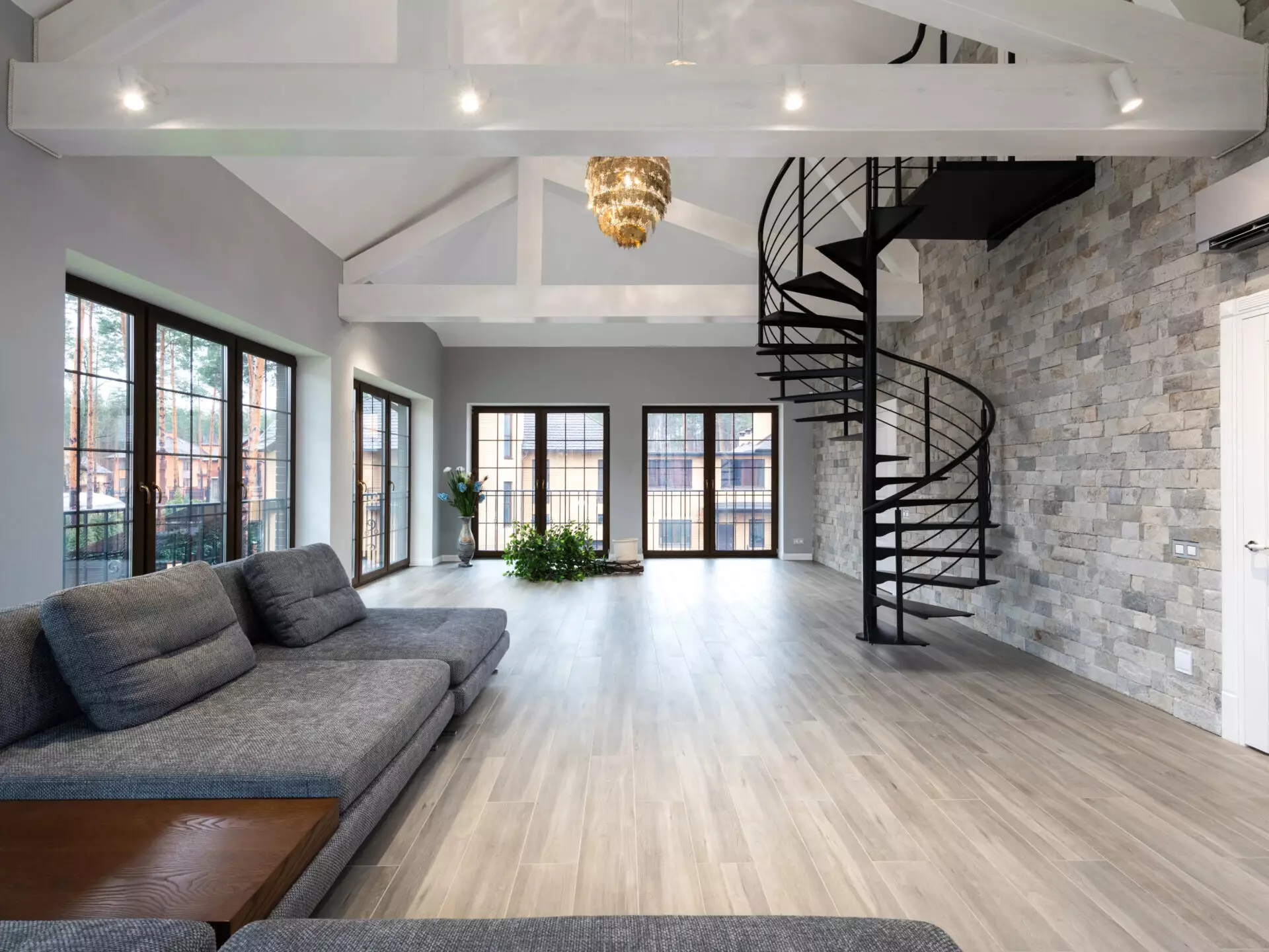 Uptown Cedar – A spacious, modern living area featuring vaulted ceilings with exposed white beams, light wood-look flooring, and a textured stone accent wall. A sleek black metal spiral staircase leads to an upper level, complemented by a large grey sectional sofa and multiple dark-framed glass doors that bring in natural light.