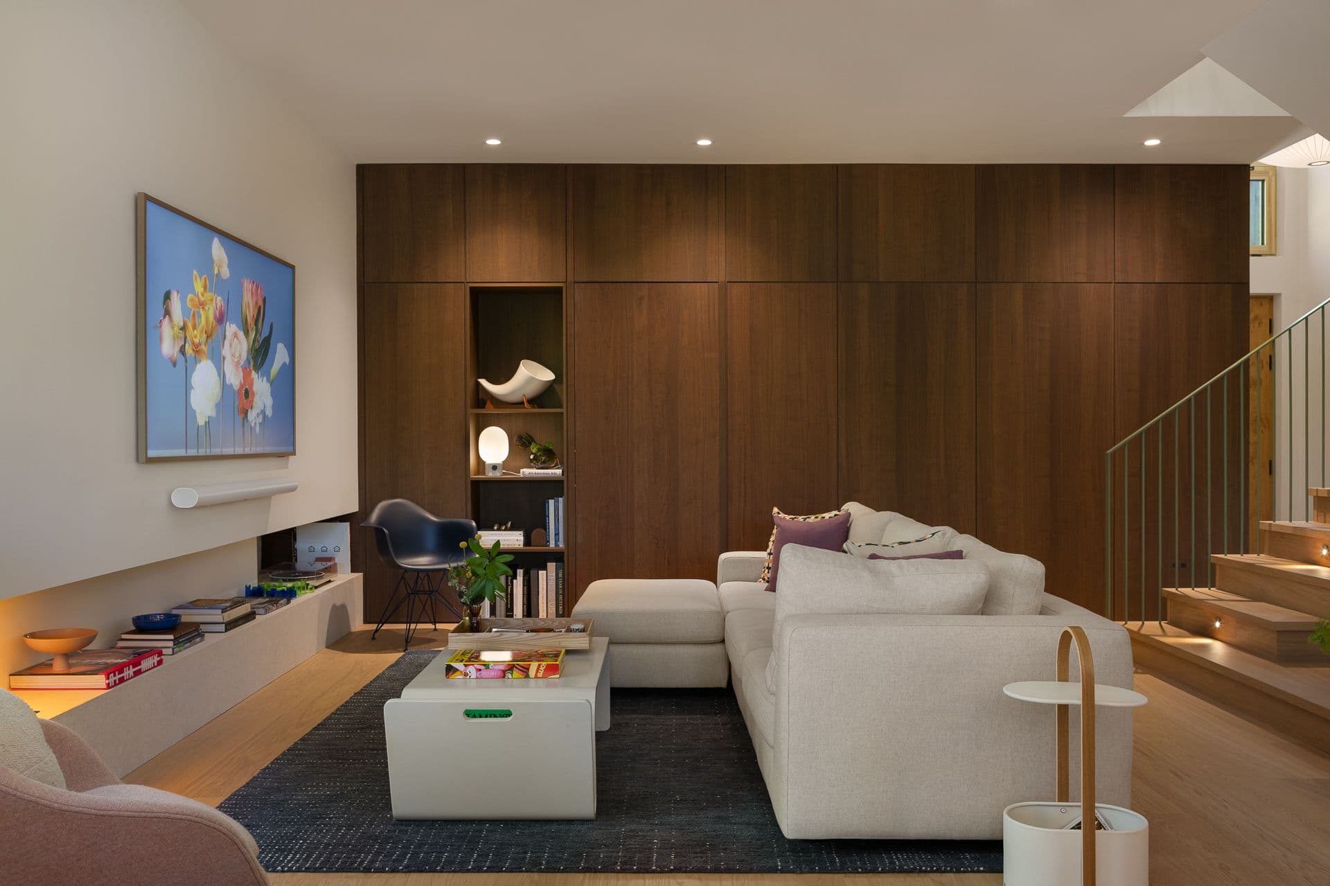 Ebor Plateau – A modern living room features a large beige sectional sofa arranged around a low coffee table atop a dark area rug. Full-height dark wood paneling with integrated shelving lines the back wall, complemented by light hardwood floors, recessed ceiling lights, and an adjacent staircase with open wooden treads.