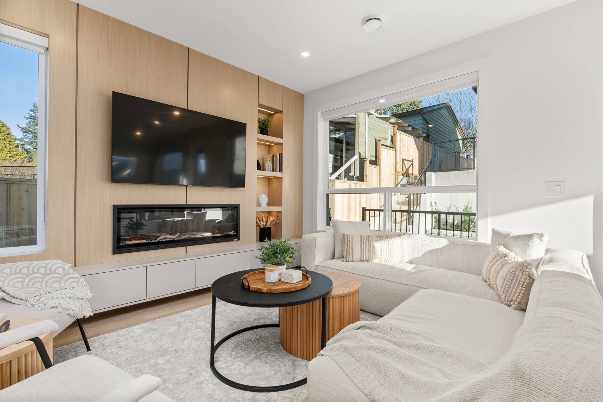 Ebor Plateau – A bright, contemporary living room features a custom light-wood feature wall with a mounted flat-screen TV, linear electric fireplace, and backlit open shelving. A plush white sectional sofa and nesting coffee tables rest on a light patterned rug, illuminated by natural sunlight streaming through large windows.