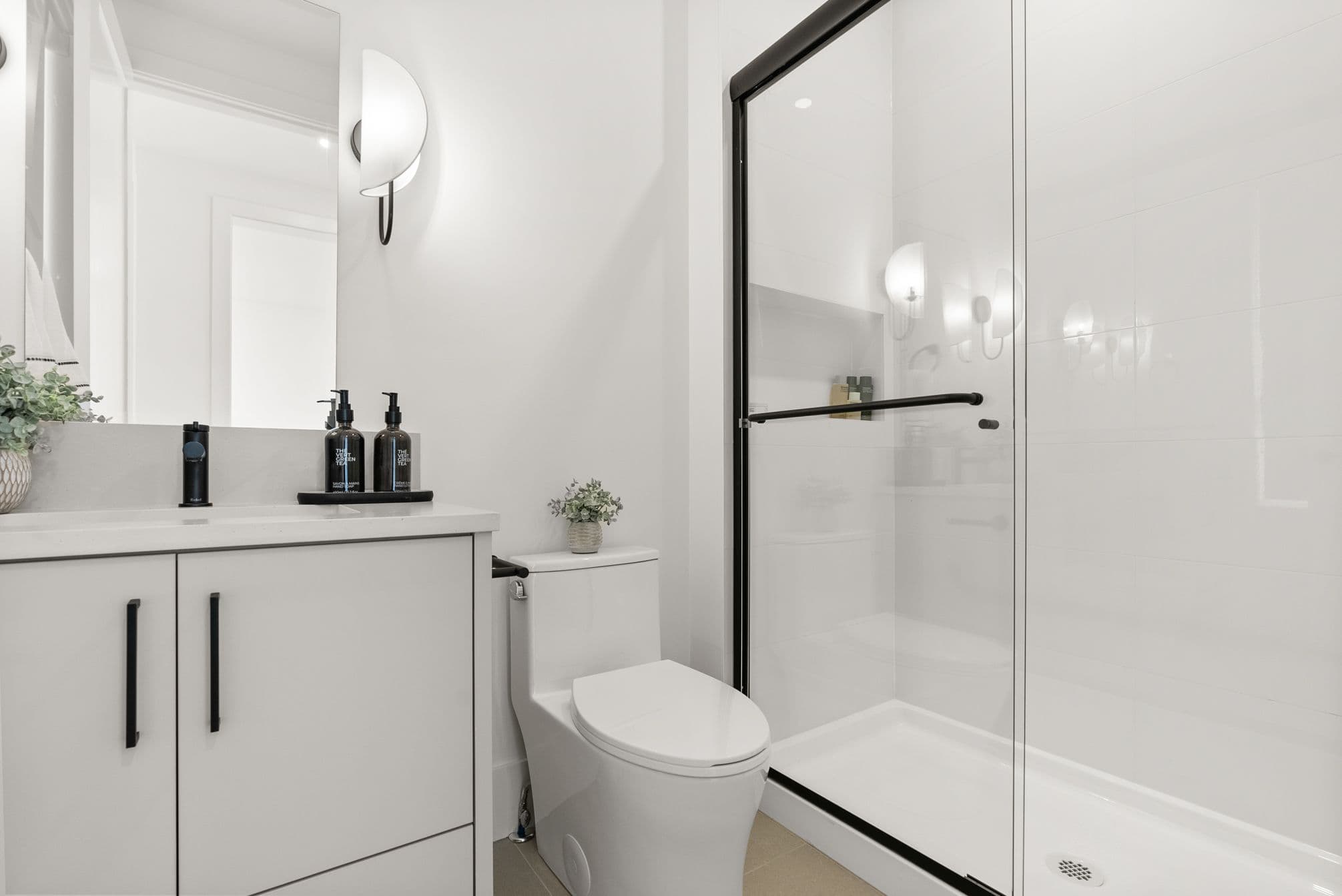 Ebor Plateau – A bright, modern bathroom featuring a white vanity with black hardware, a matching toilet, and a glass-enclosed walk-in shower with black framing. A sleek sconce light illuminates the white walls and large mirror, creating a clean and minimalist aesthetic.