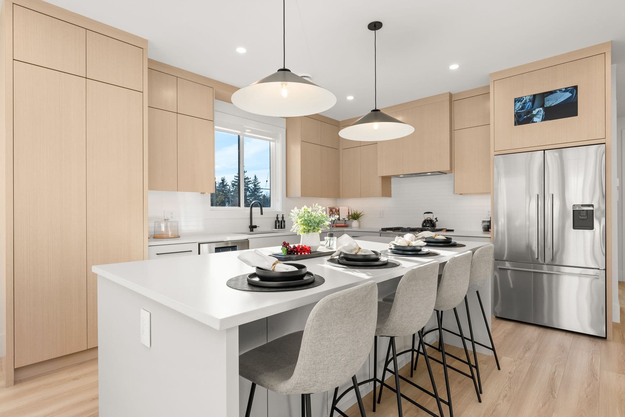 Ebor Plateau – A modern, bright kitchen featuring light wood flat-panel cabinetry, white quartz countertops, and stainless steel appliances including a French-door refrigerator. A large kitchen island with four upholstered barstools is illuminated by two wide saucer pendant lights and set with dishware.