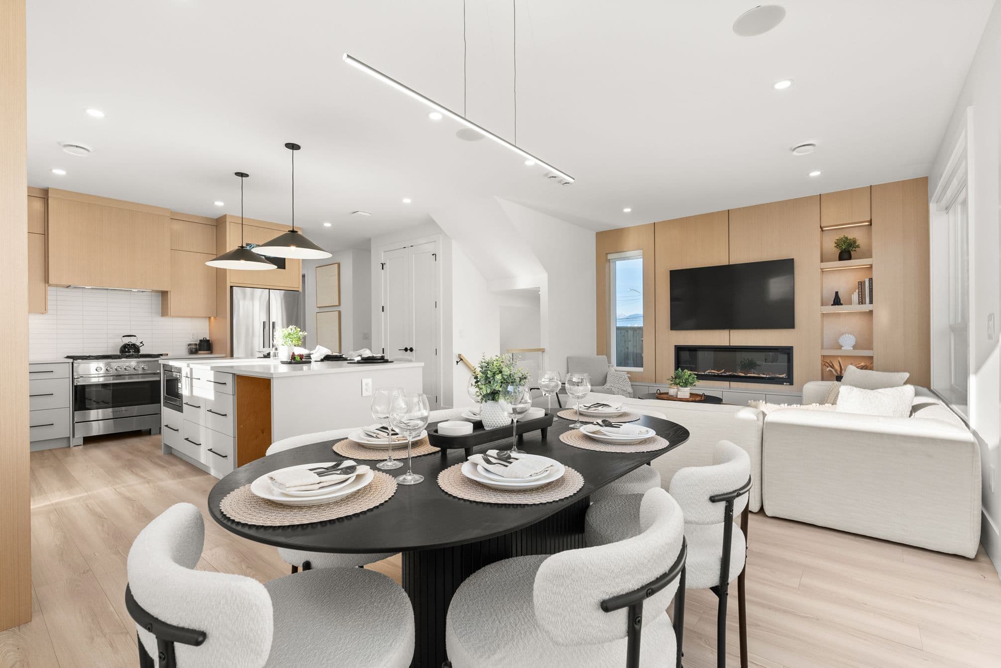 Ebor Plateau – An open-concept living, dining, and kitchen area featuring light wood floors, an oval black dining table with textured white chairs, and an adjacent living room with a built-in wood feature wall and linear fireplace. The kitchen in the background includes a waterfall island, light wood upper cabinetry, and stainless steel appliances.