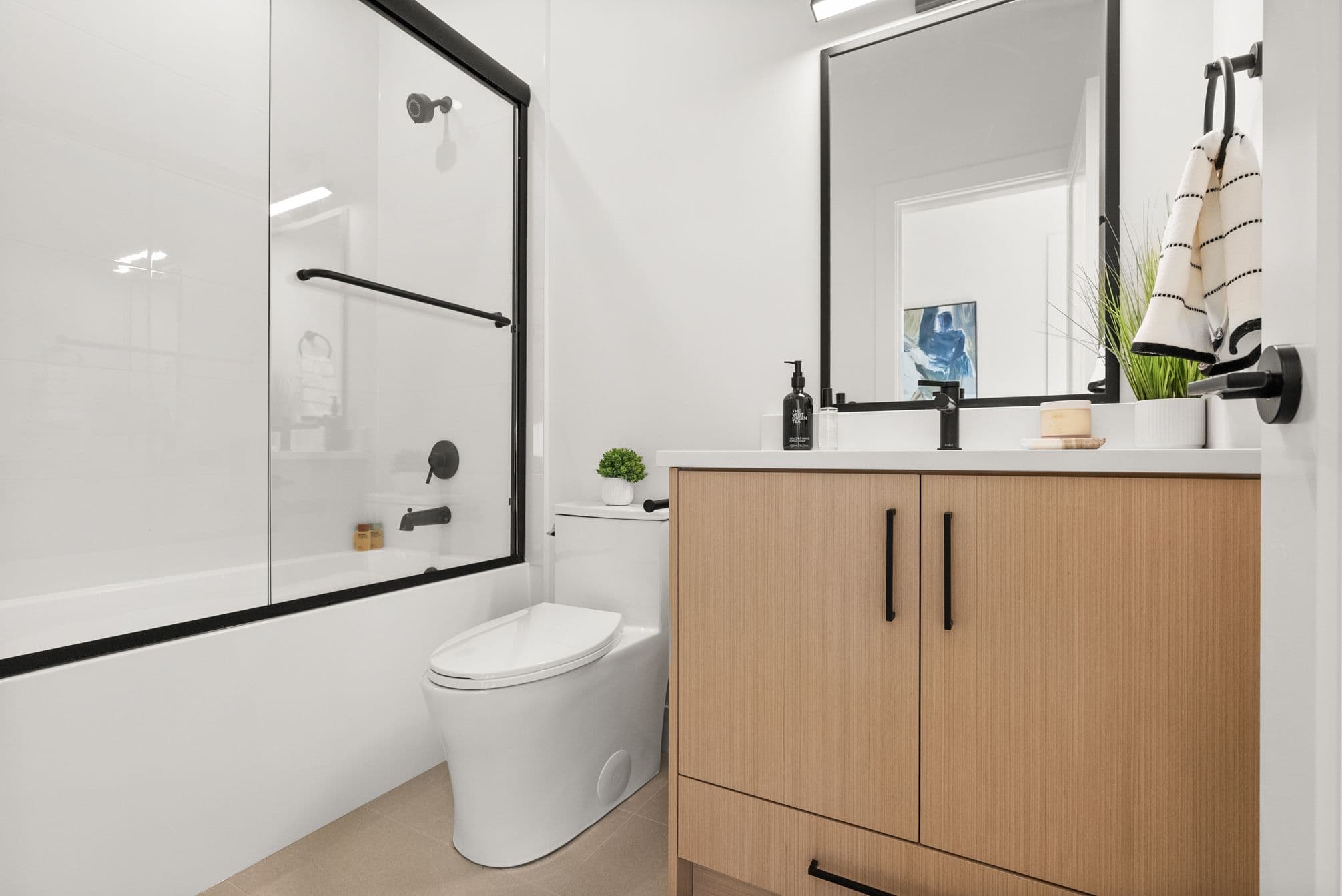 Ebor Plateau – A modern, brightly lit bathroom featuring a light wood vanity cabinet paired with matte black hardware, faucet, and mirror frame. A glass-enclosed bathtub and shower combination with black trim and fixtures sits adjacent to a sleek white toilet against crisp white walls.
