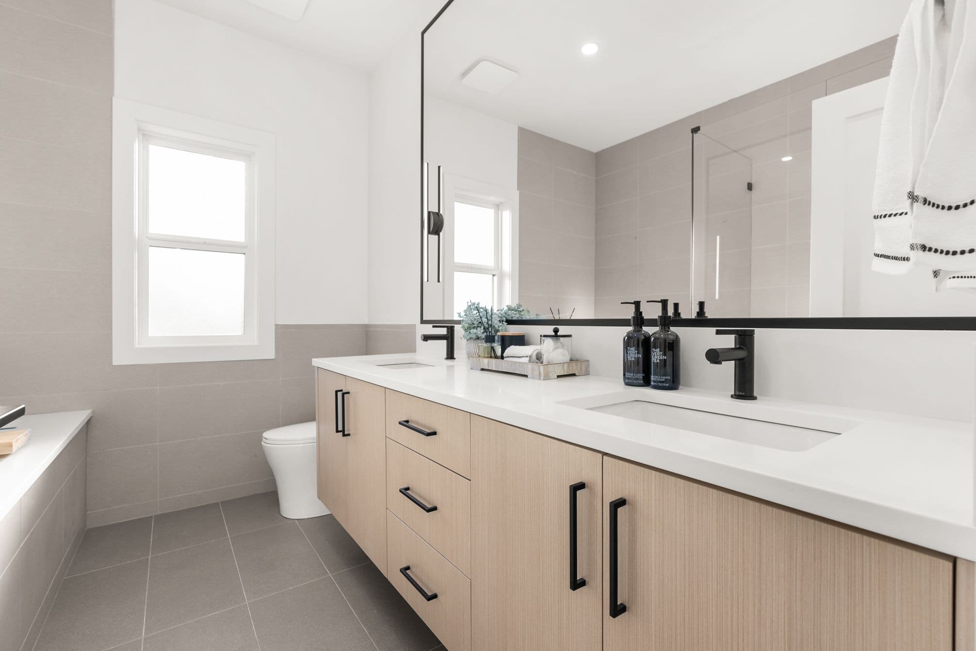 Ebor Plateau – A bright, contemporary bathroom featuring a light wood floating double vanity with a white countertop and matte black fixtures. Neutral grey tiling covers the floor and lower walls, while a large black-framed mirror reflects a glass-enclosed walk-in shower.