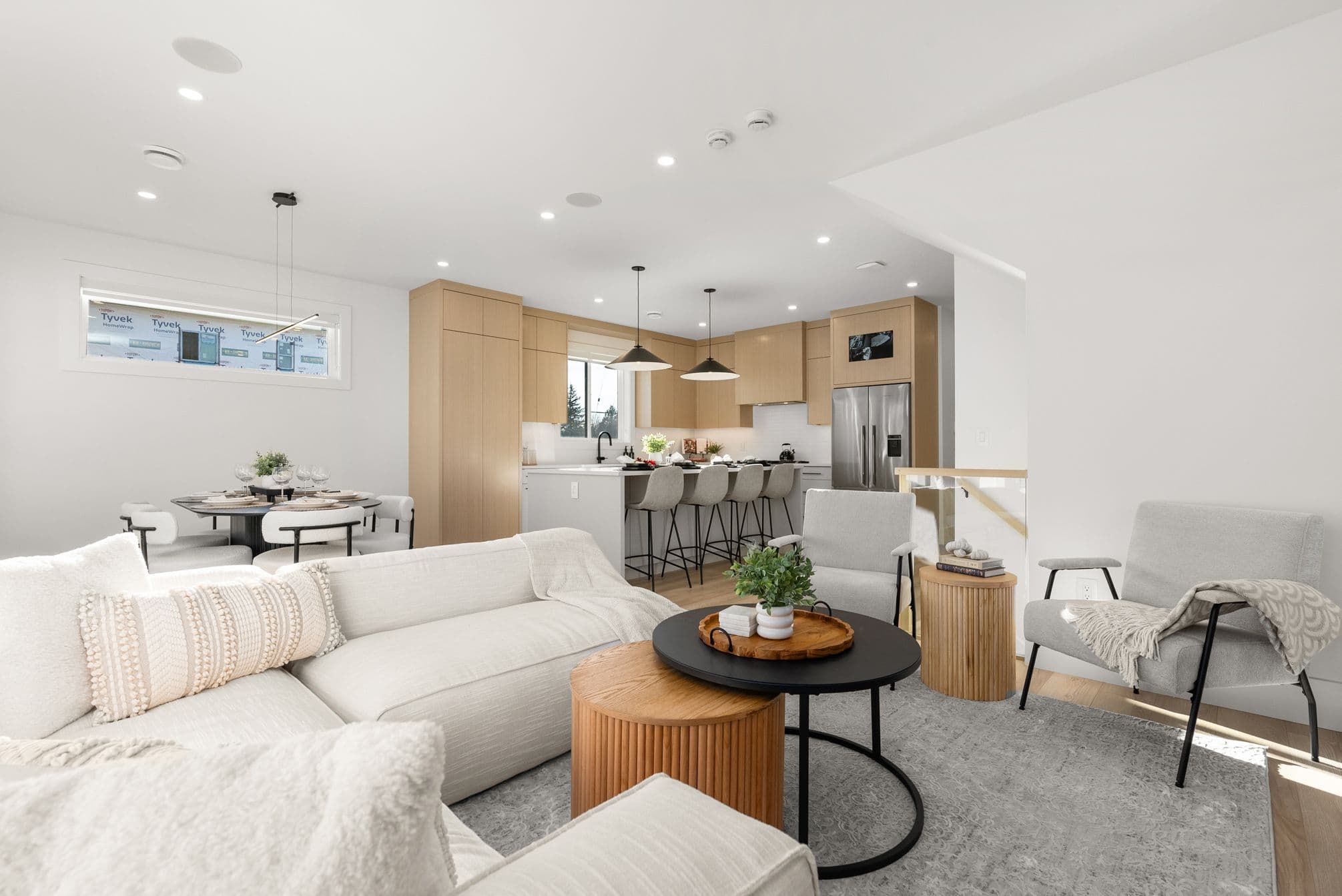 Ebor Plateau – A bright, open-concept living space features an off-white sectional and armchairs centered around wooden and black round coffee tables, flowing seamlessly into a kitchen with light wood cabinetry, stainless steel appliances, and a breakfast bar island. Recessed ceiling lighting and pendant fixtures illuminate the clean, contemporary interior, which also includes a small dining area adjacent to the
