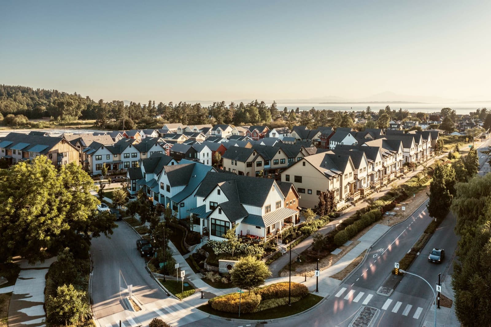 Southlands Tsawwassen – Aerial view at sunset of a master-planned neighborhood of houses and townhomes with streets, sidewalks, and trees, with the ocean and mountains visible in the distance.