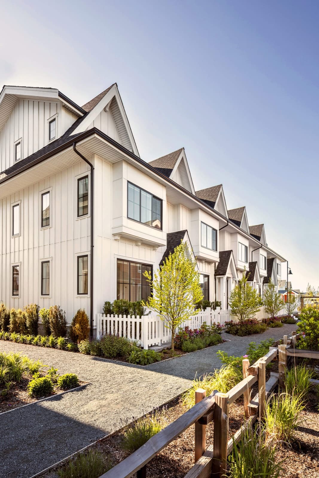 Southlands Tsawwassen – Row of white board-and-batten townhomes with steep gabled roofs, small front gardens, and white picket fences along a gravel walkway.