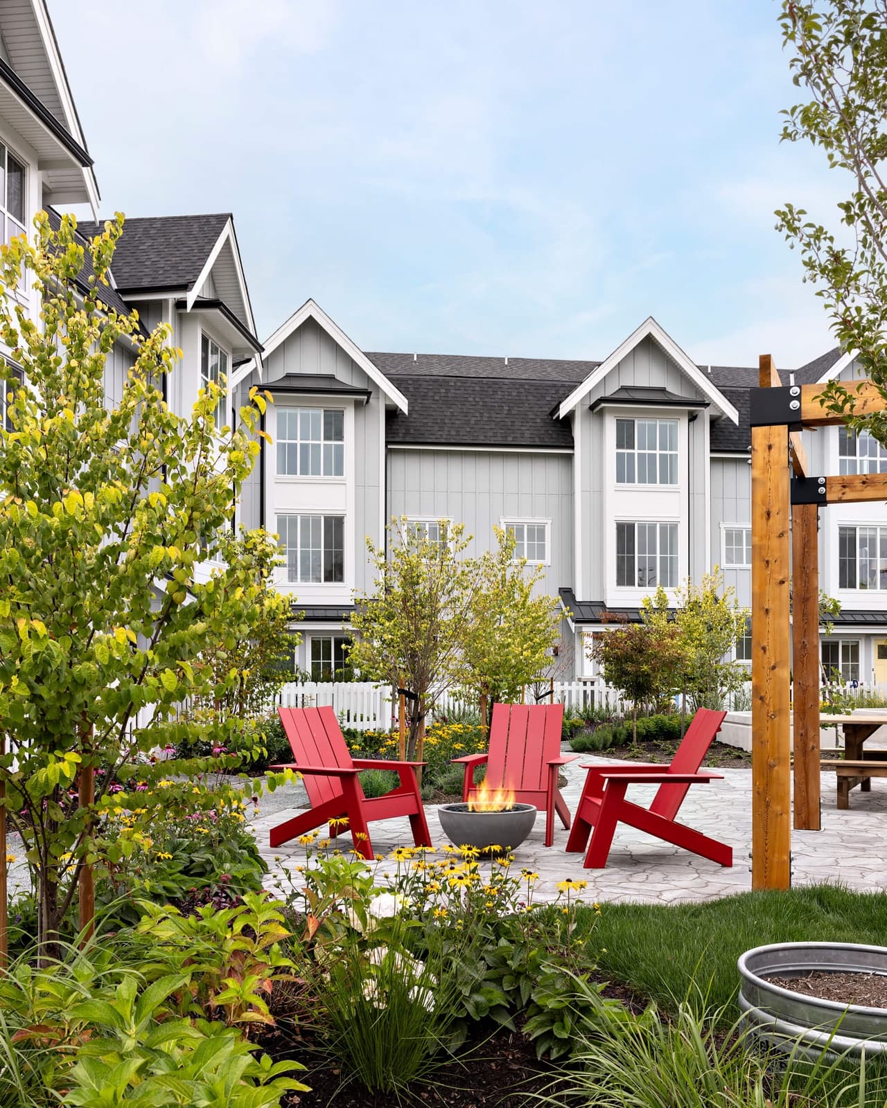 Southlands Tsawwassen – Outdoor gathering area with three red adirondack chairs around a lit fire bowl on a stone patio, in front of grey townhomes with white trim.