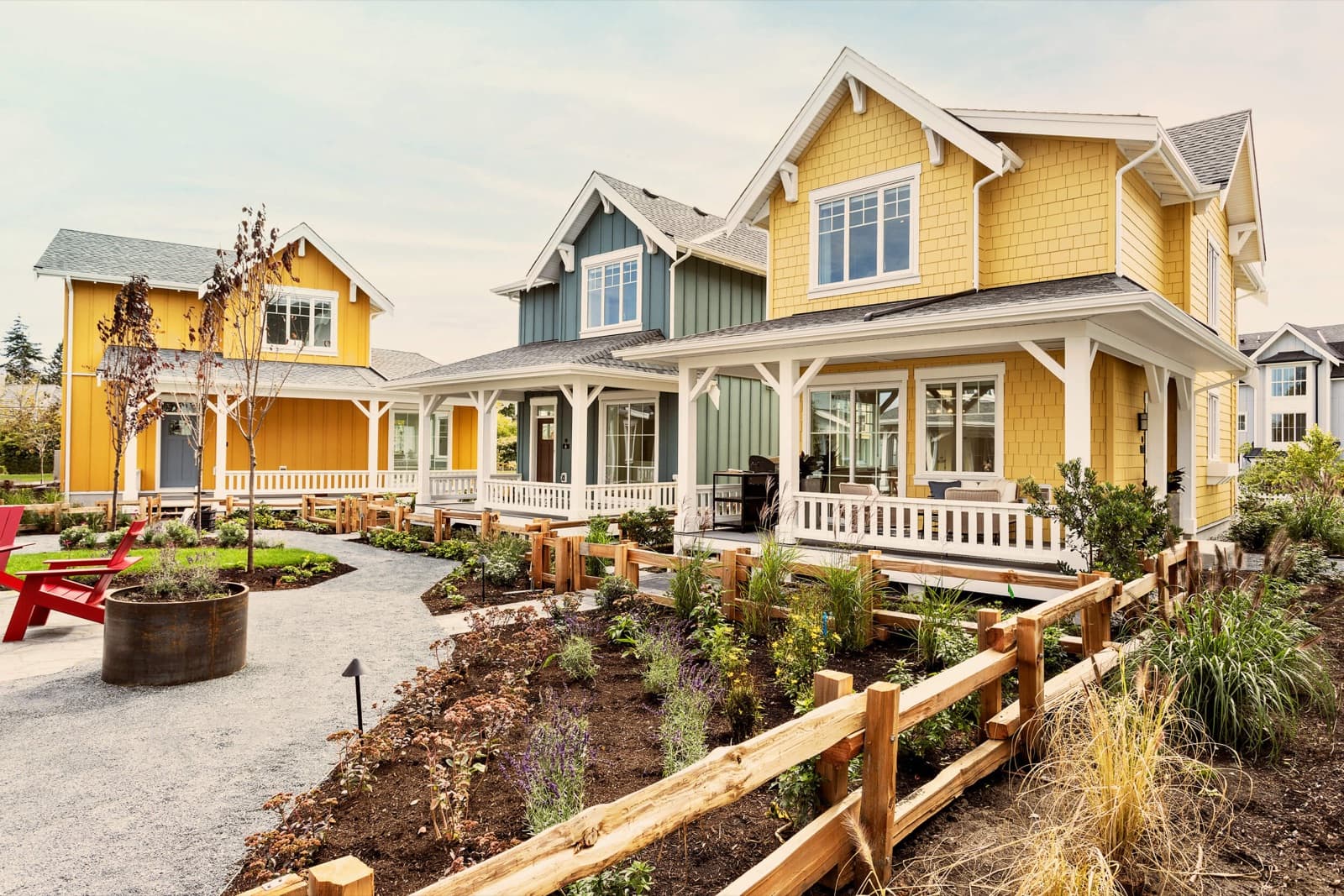 Southlands Tsawwassen – Row of craftsman-style cottage homes in yellow and blue-grey with white wraparound porches, gabled roofs, and a red adirondack chair on the front lawn.
