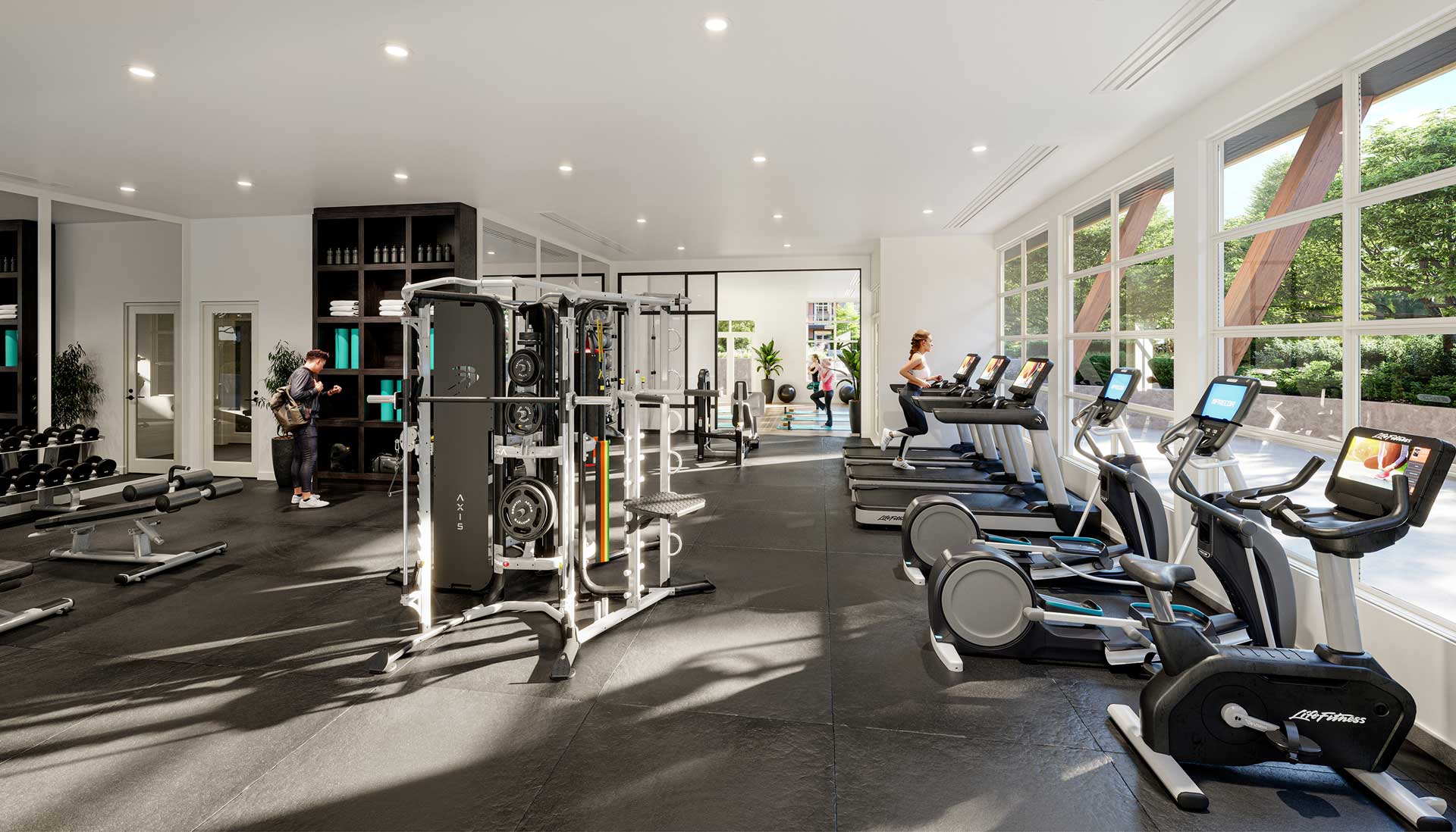 Inspire – Bright shared fitness room with a weight rack, dumbbells, treadmills, ellipticals, and large windows looking outside.