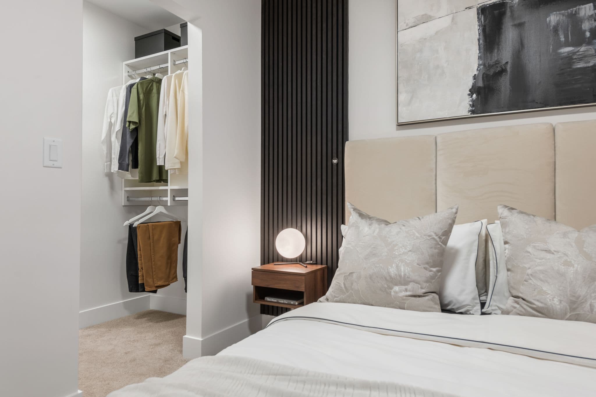 Aria 2 – Bedroom with a beige upholstered headboard, abstract wall art, a nightstand lamp, and a walk-in closet visible through the doorway.