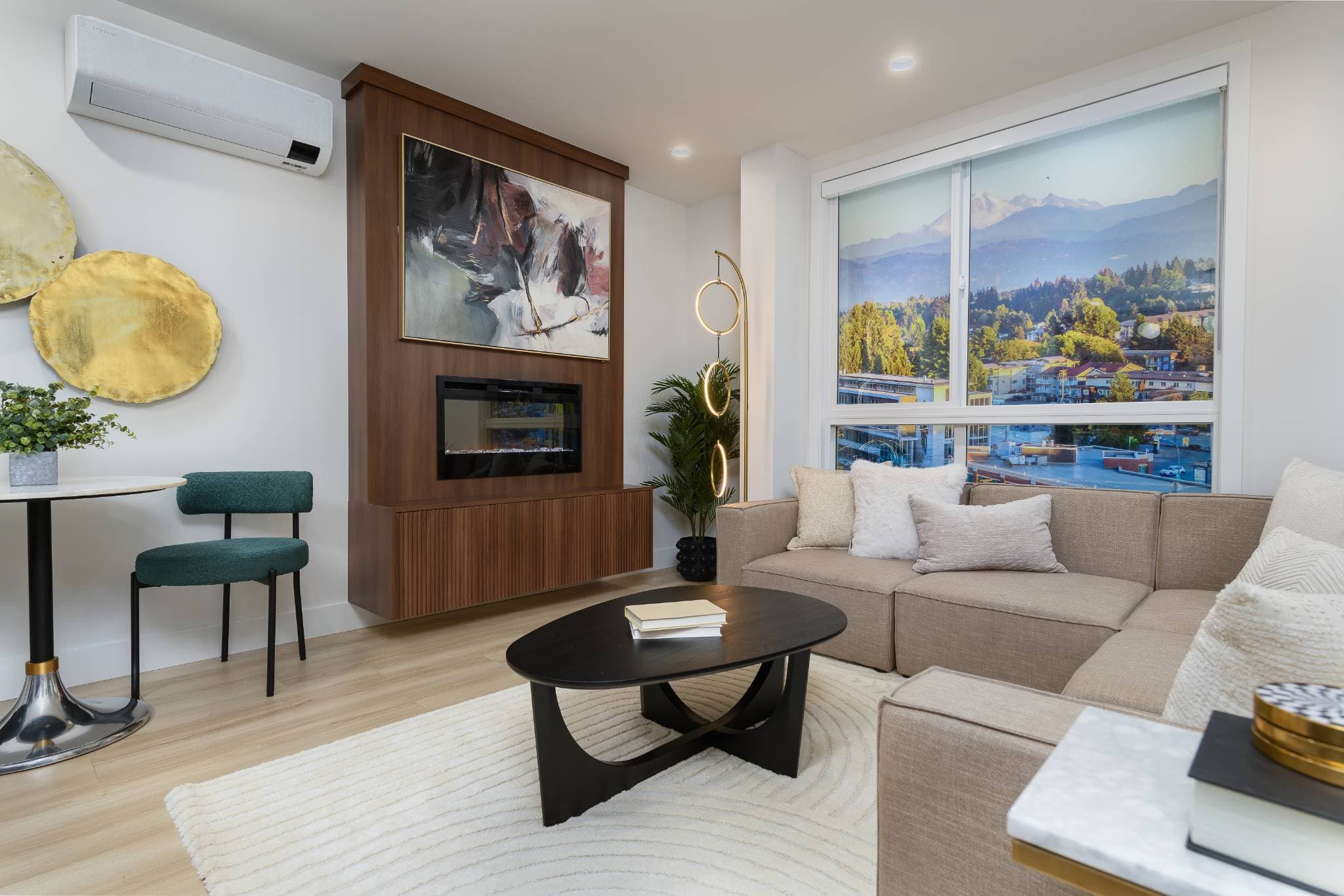 Aria 2 – Living room with a built-in fireplace, abstract art panel, beige sectional sofa, and a window showing a mountain view.
