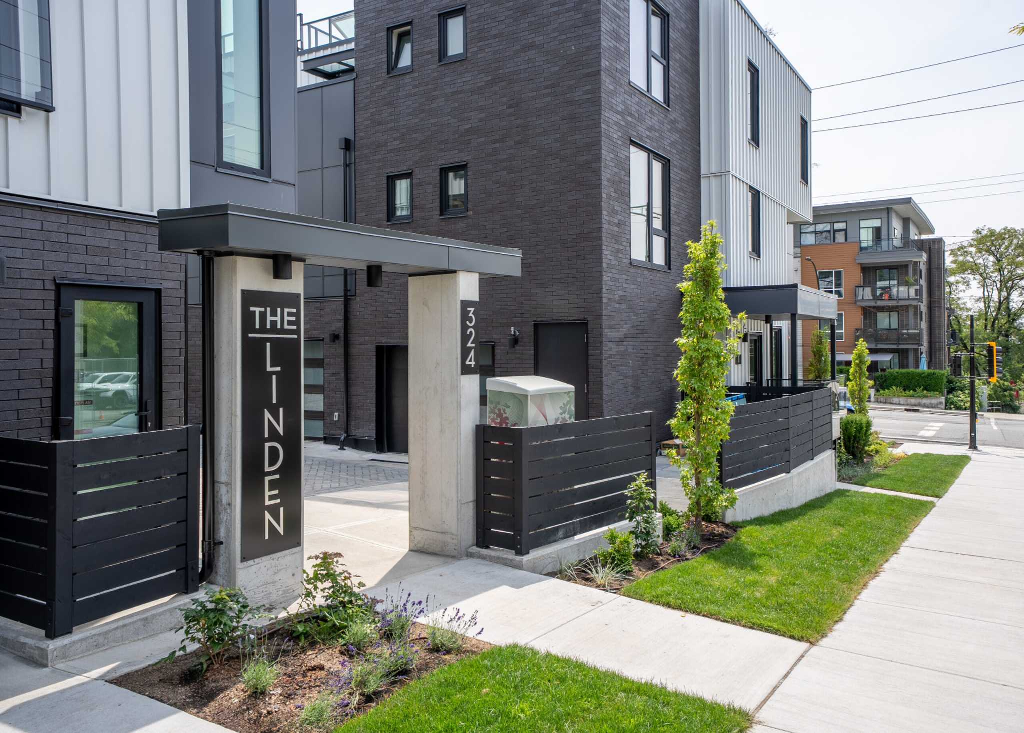 The Linden Townhomes – Street-level view of a dark brick townhome entrance with a sign reading THE LINDEN, unit number 324, a black slat fence, and a landscaped walkway with young trees.