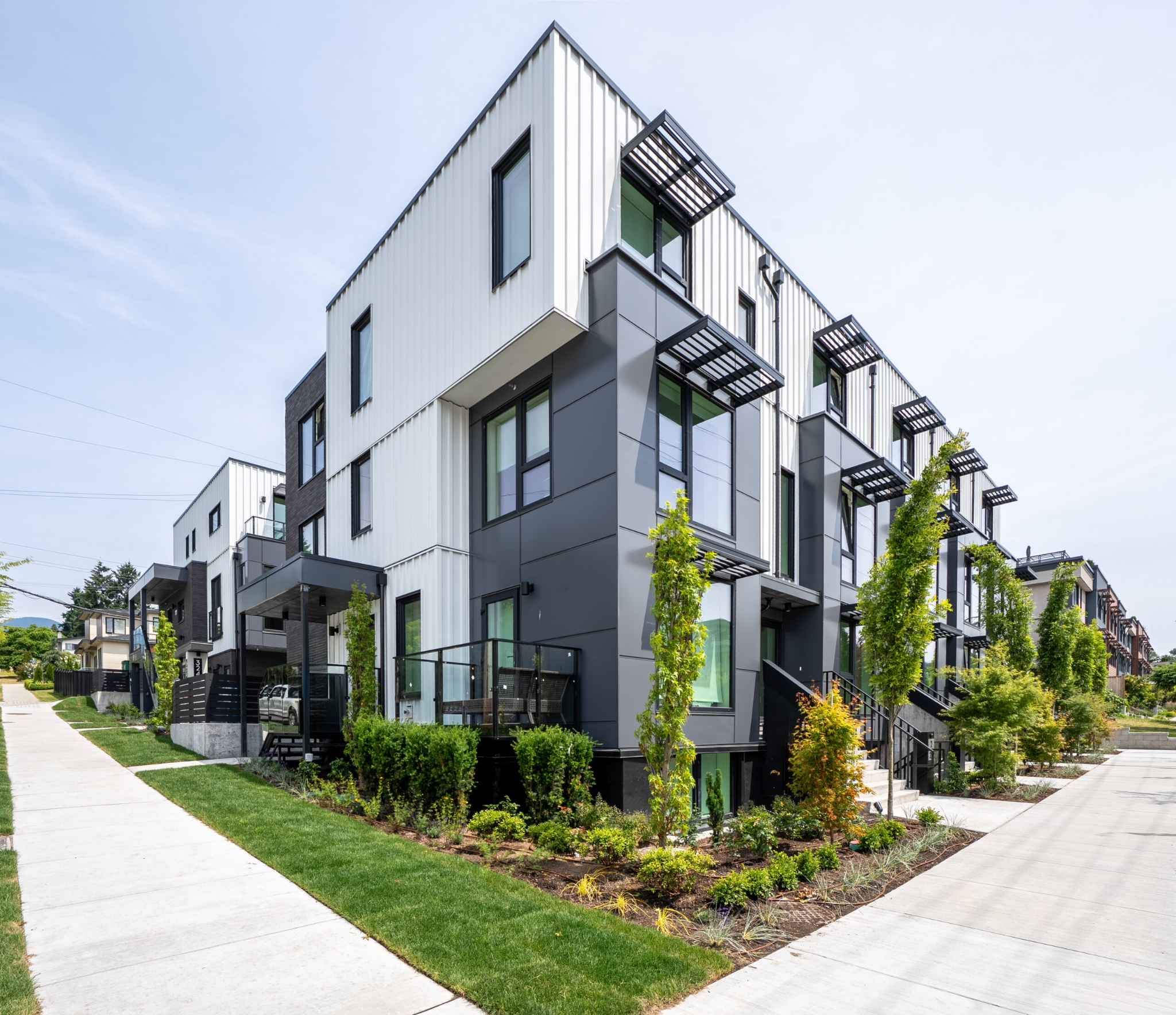 The Linden Townhomes – Corner view of a modern multi-unit townhome building with dark and white metal and brick cladding, black-framed windows, balconies, and a row of young trees along the sidewalk.