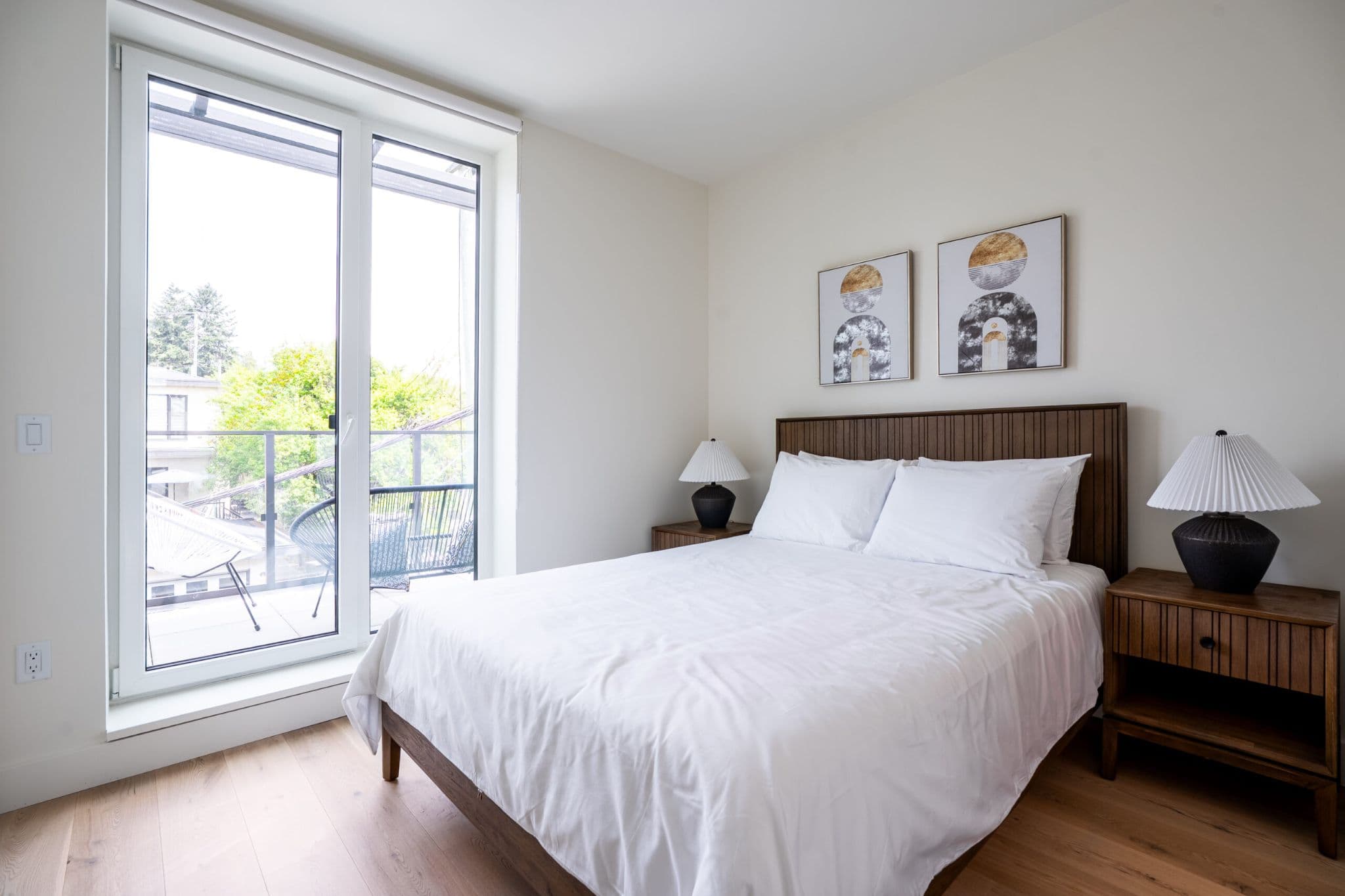 The Linden Townhomes – A bedroom with a queen bed dressed in white linens, a wood headboard, matching nightstands and lamps, and a glass door leading out to a small balcony with outdoor seating.
